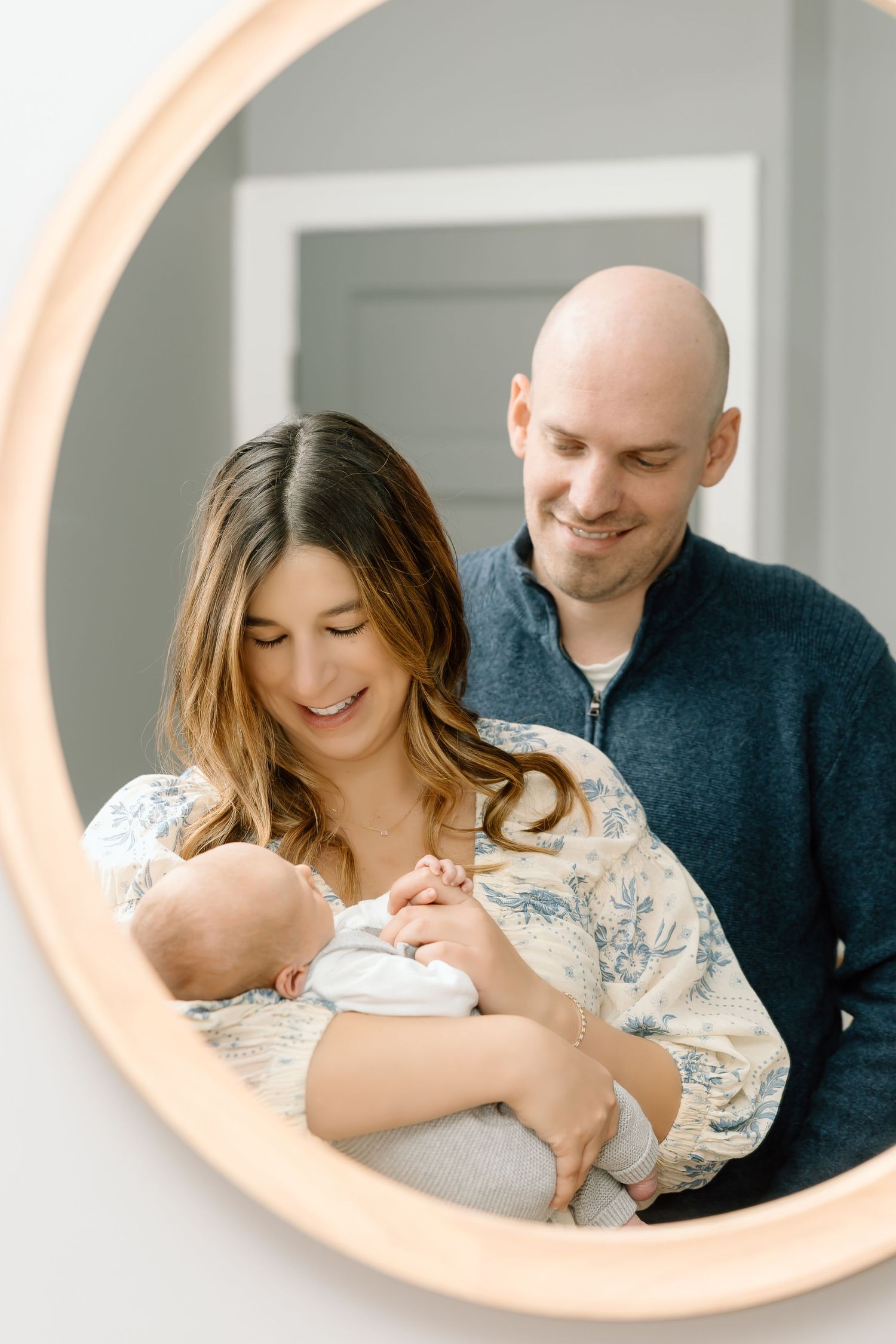 A couple shares a tender moment with their newborn baby while looking in a round mirror with a peach-colored frame.