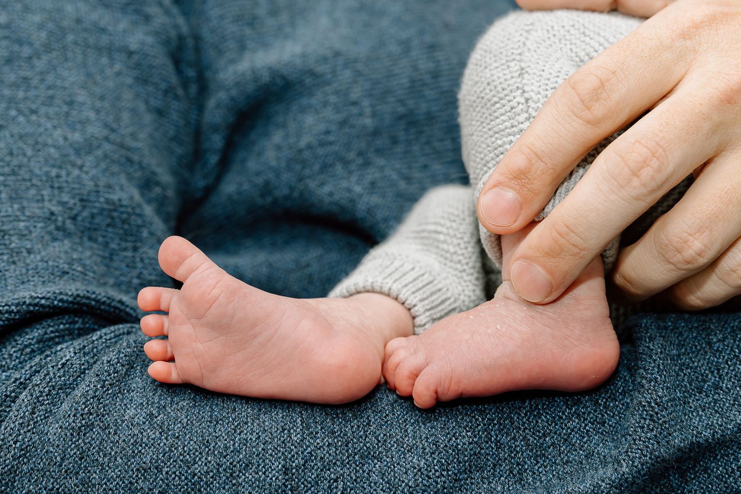 Close-up of tiny baby feet in white socks being gently touched by adult hands on dark blue fabric.