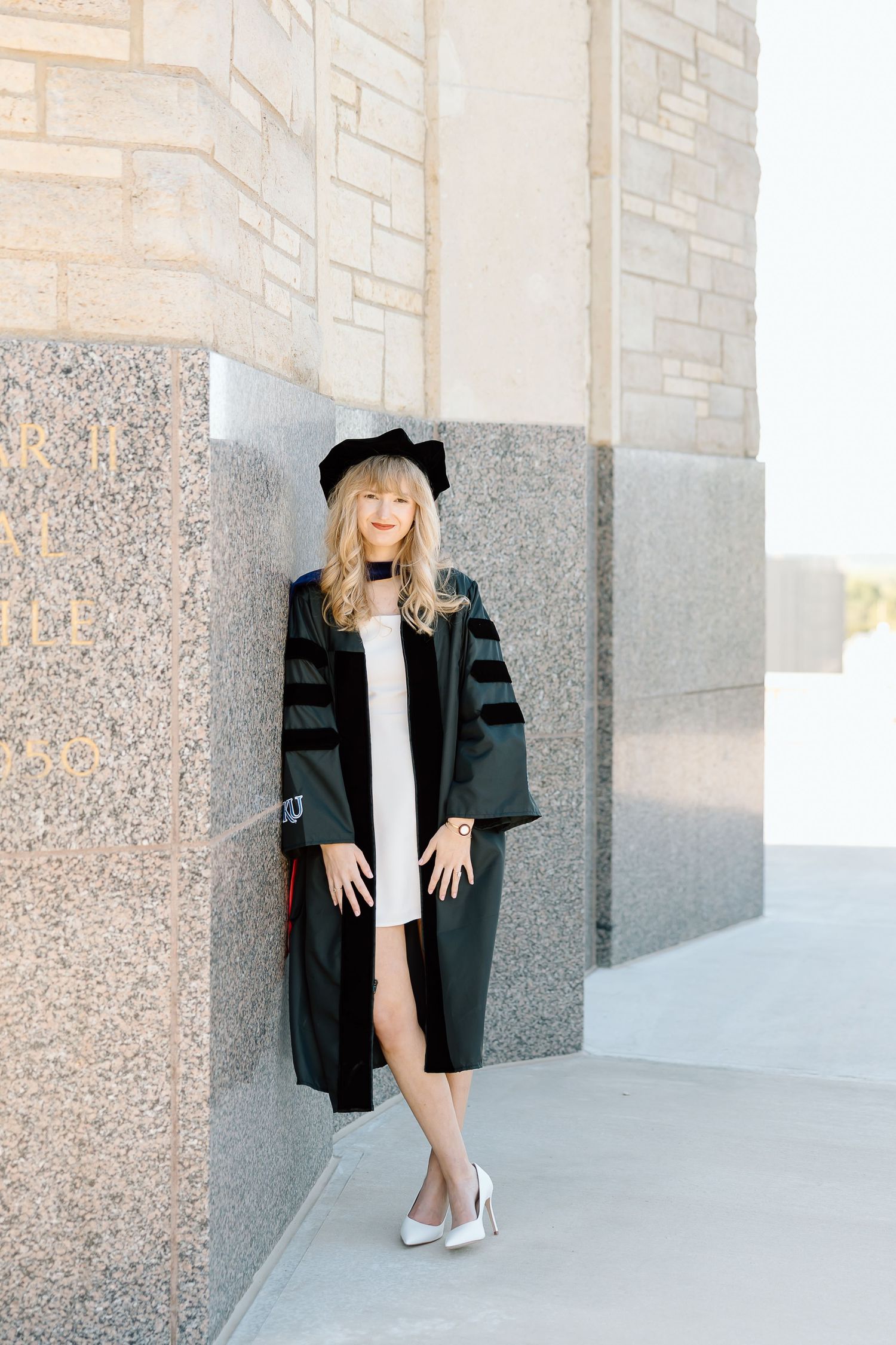 A person wearing a black blazer and light dress poses against a textured concrete wall in an urban setting.