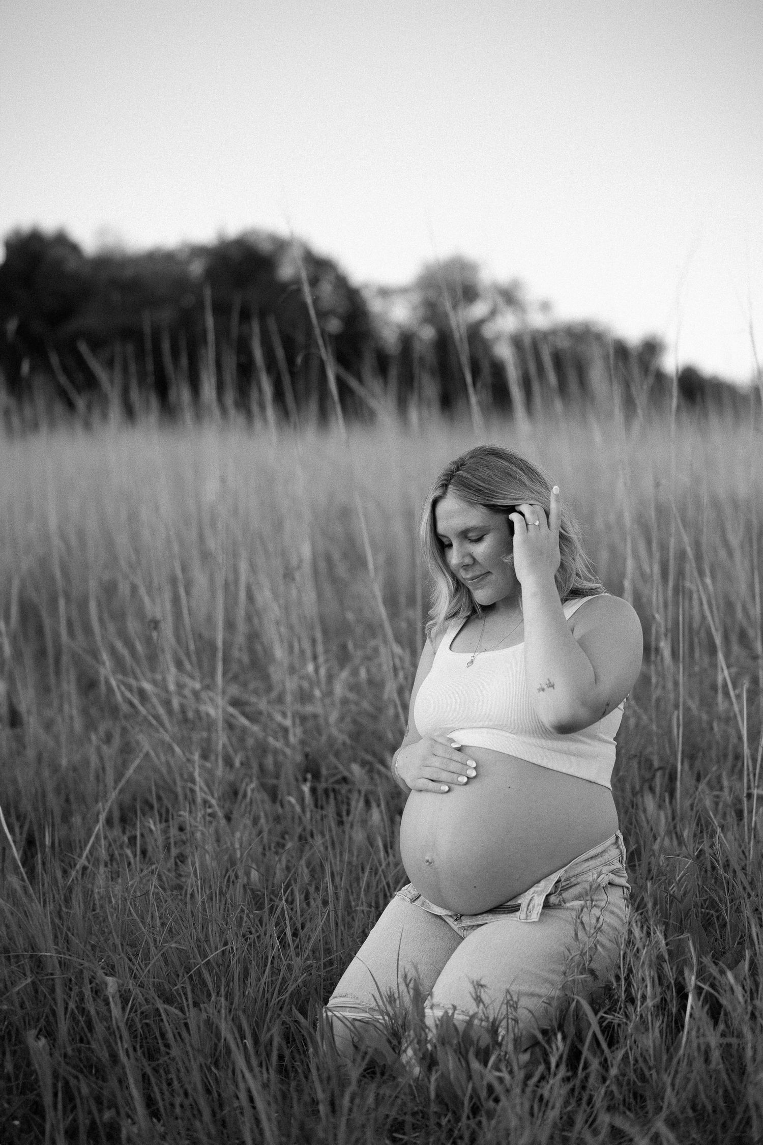 A black and white maternity portrait shows an expectant mother sitting thoughtfully in a field of tall grass at sunset.