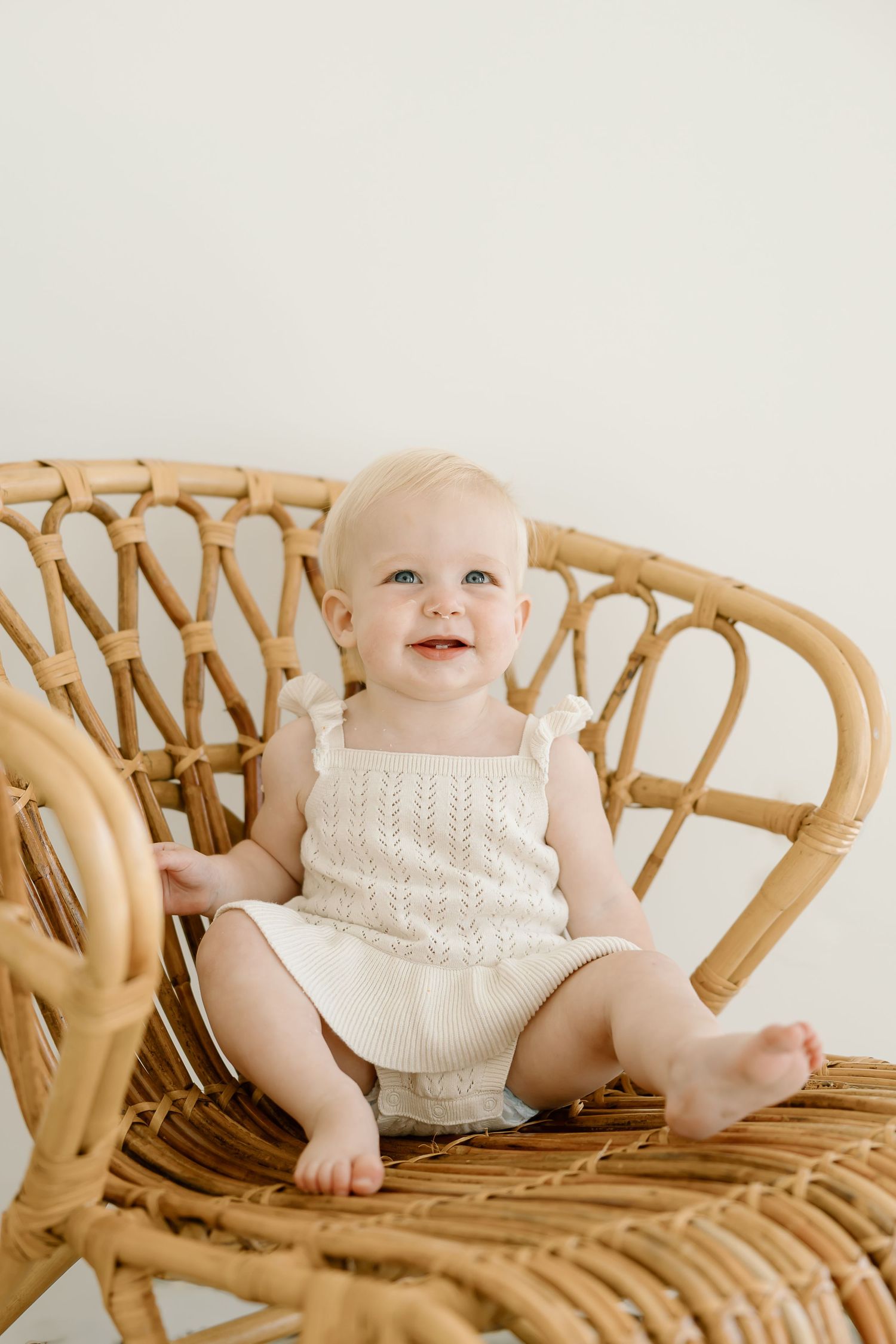 A baby wearing a white knit sweater sits and smiles on a vintage rattan bench in a series of candid poses.