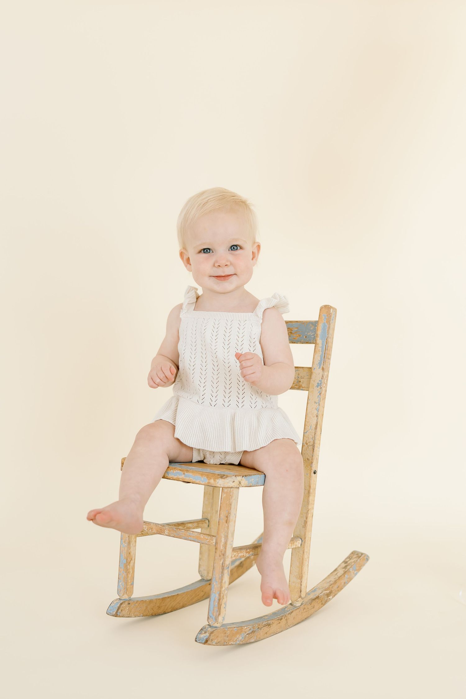 A toddler in white clothing sits on a wooden rocking chair against a cream background.