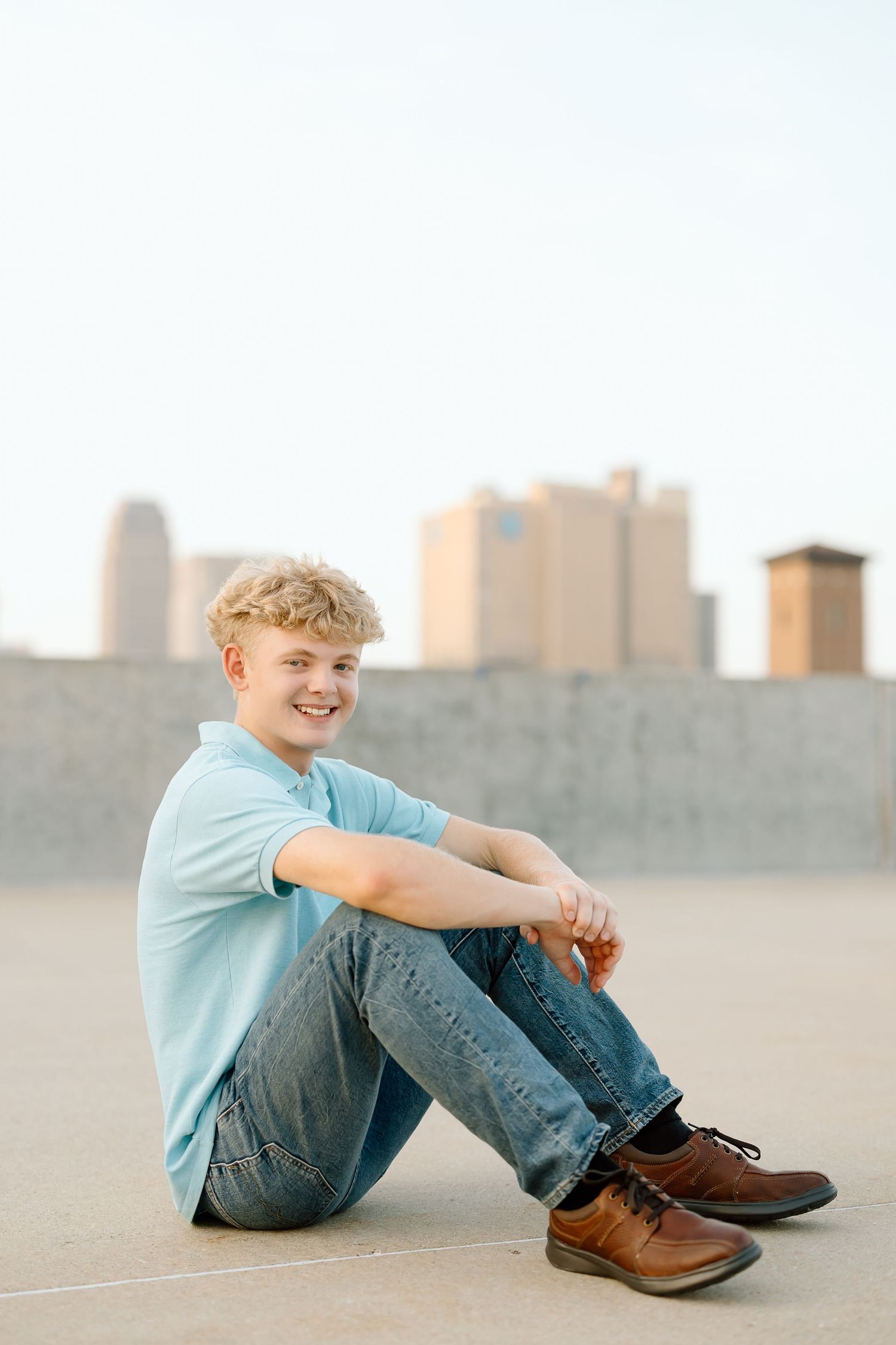 A person in a light blue shirt and jeans sits casually on a rooftop with city buildings visible in the background.