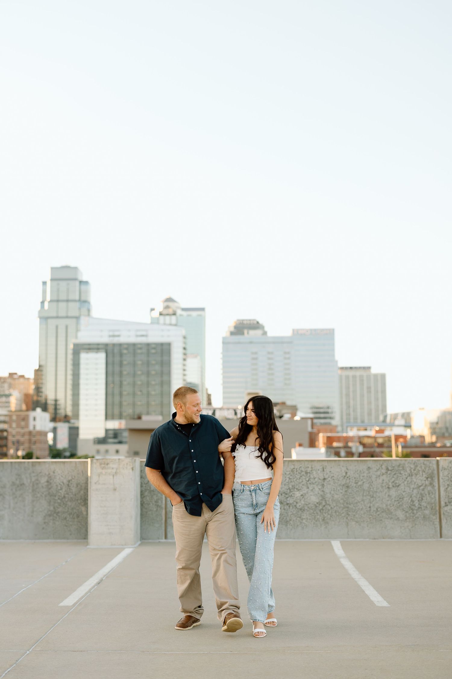 A couple sits together on a rooftop parking garage with city buildings in the background during golden hour.