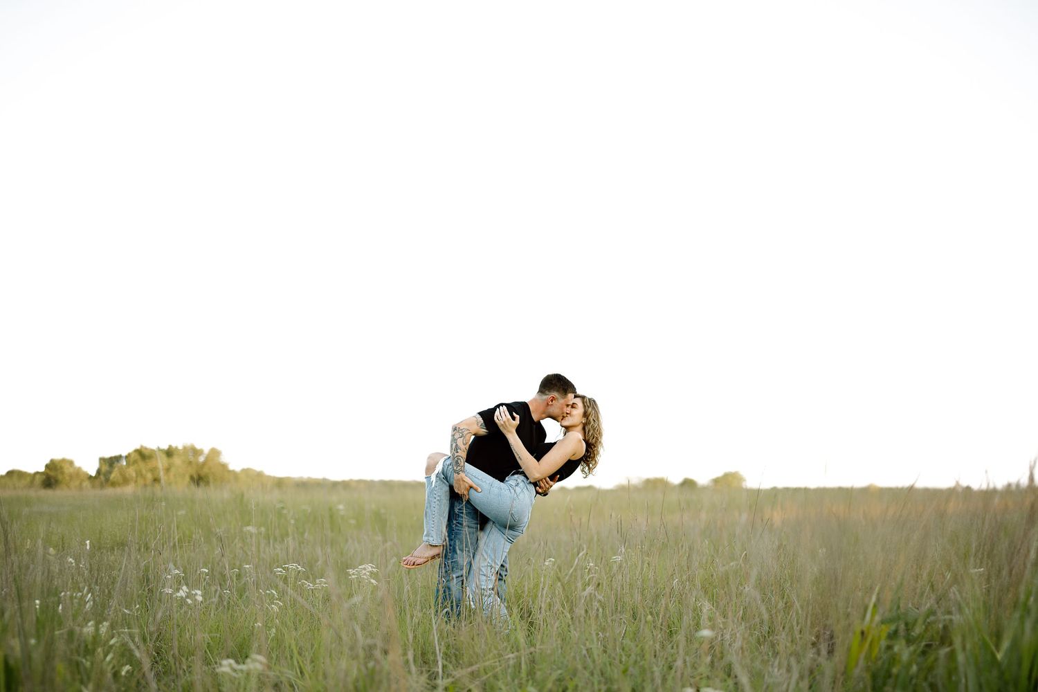 A couple embraces in a sunlit meadow with tall grass and wildflowers during a romantic outdoor photoshoot.