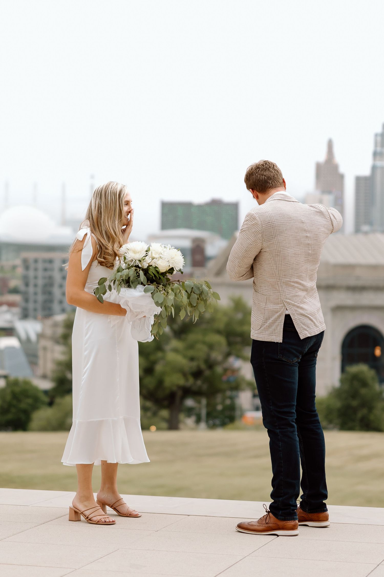 A couple in wedding attire stands together holding flowers against an urban skyline background.