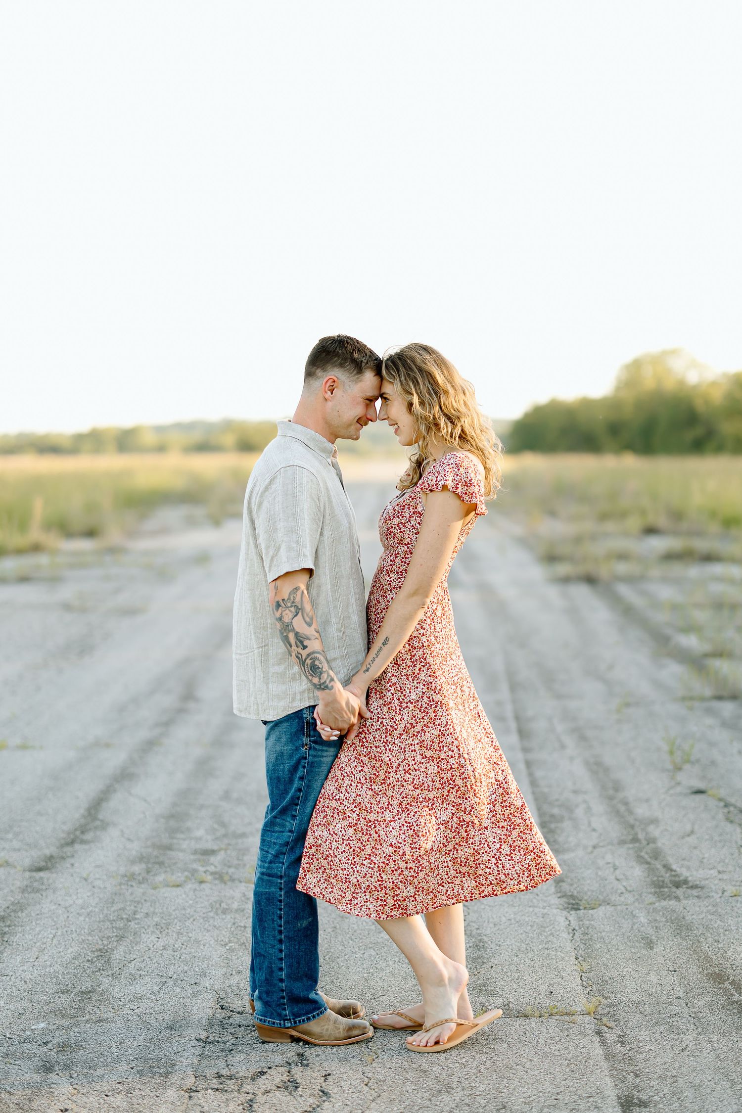 A romantic couple stands close together on a dirt road at sunset, with her wearing a pink floral dress and him in casual attire.