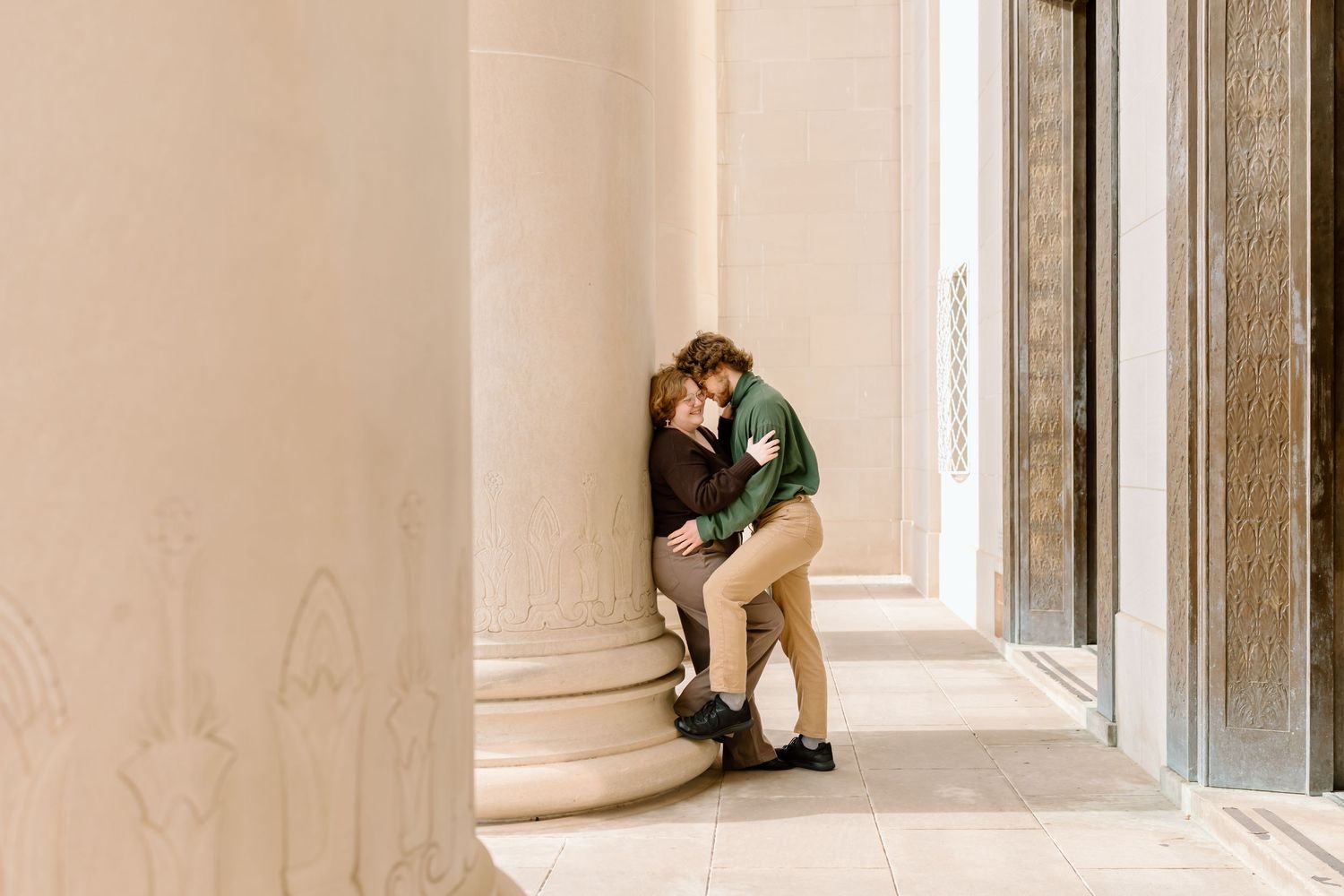 A person in a green sweater sits alone in a corner of a beige hallway with natural lighting.