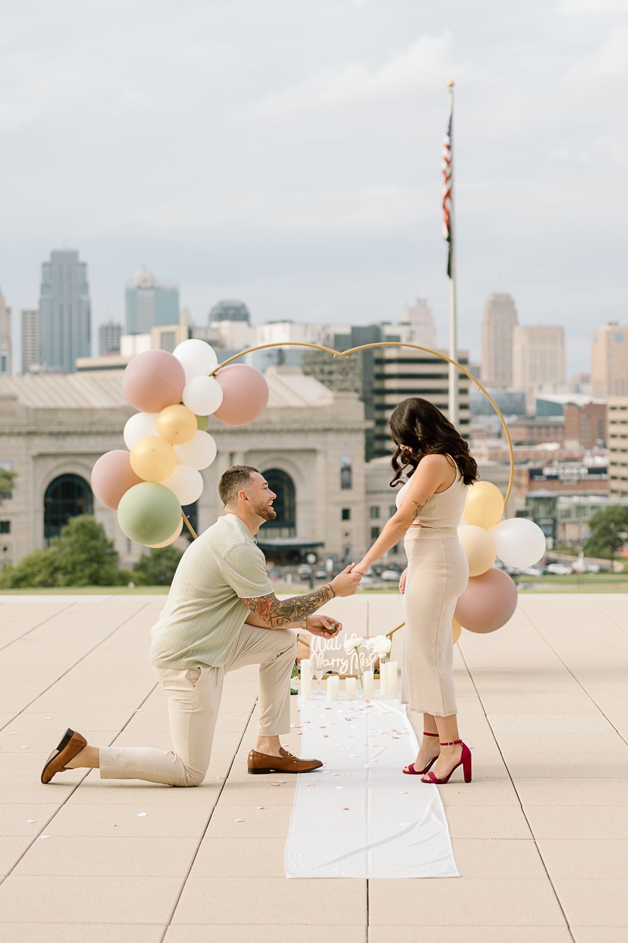 Romantic rooftop proposal scene with pink balloons and Kansas City skyline in background.