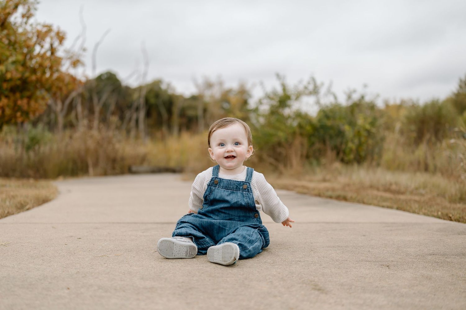 Small child in denim overalls sits on a concrete path surrounded by autumn foliage.