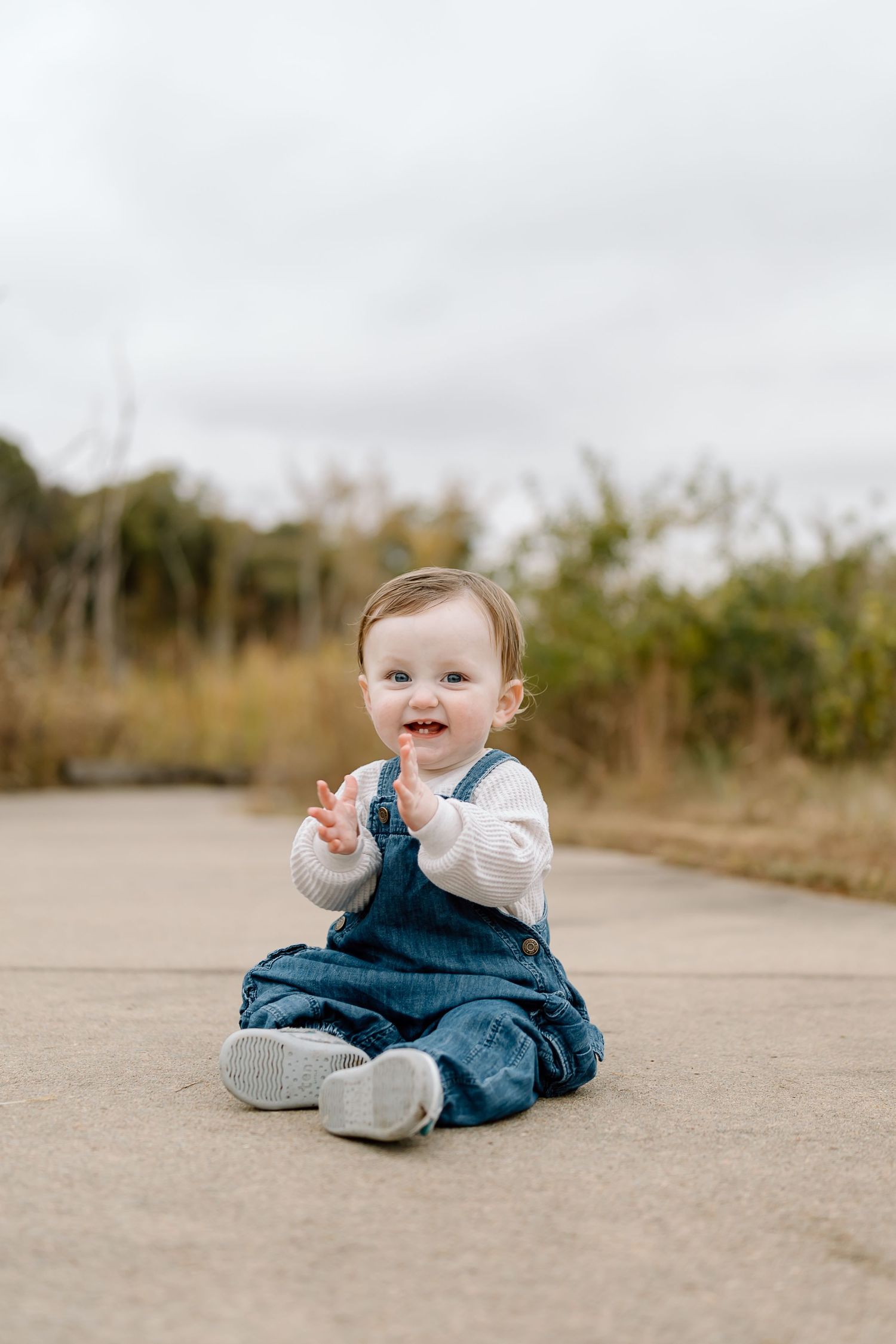 Happy baby wearing denim overalls and white shirt sits on concrete path outdoors on autumn day.