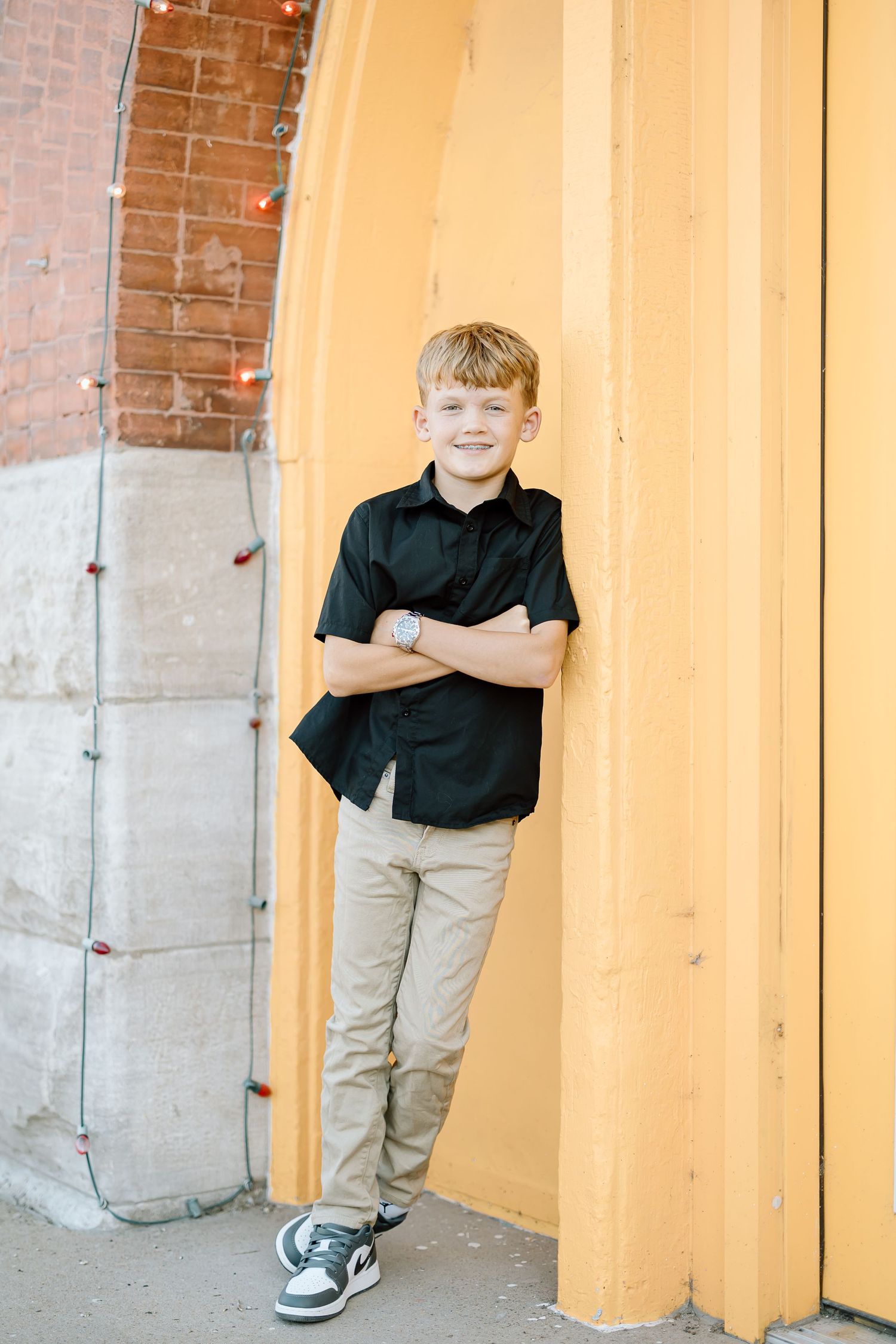 A child in a black polo shirt and khaki pants poses playfully against an orange and brick wall.