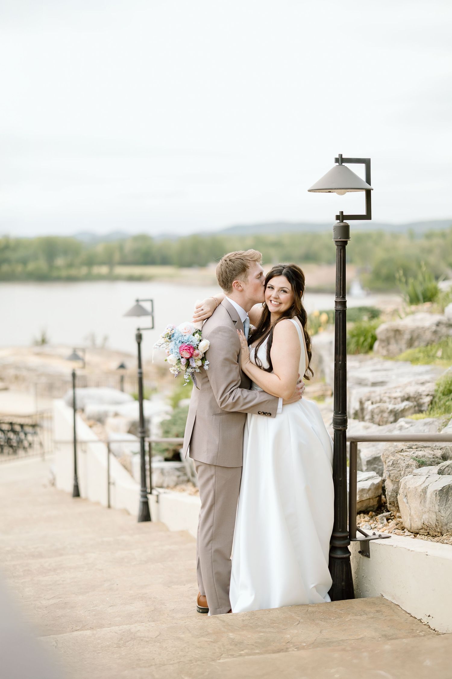 A romantic wedding couple shares tender moments at a scenic overlook with stone walls and lamp posts at sunset.