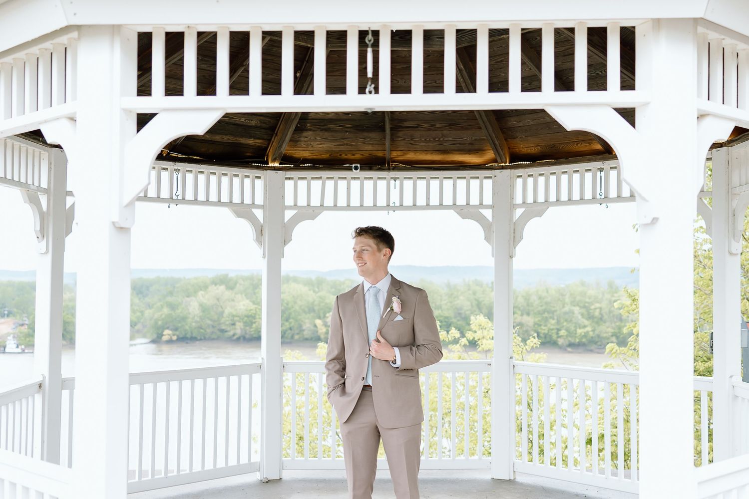 A person in a tan suit stands alone in an elegant white gazebo overlooking a scenic landscape.