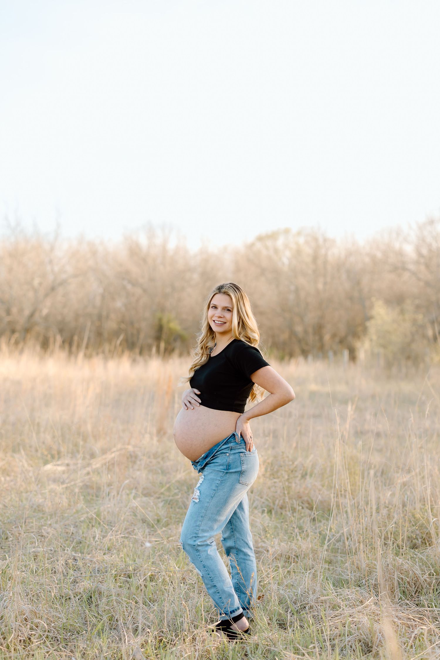 Expectant mother in black top and jeans poses in a sunlit field with tall grass and trees in the background.