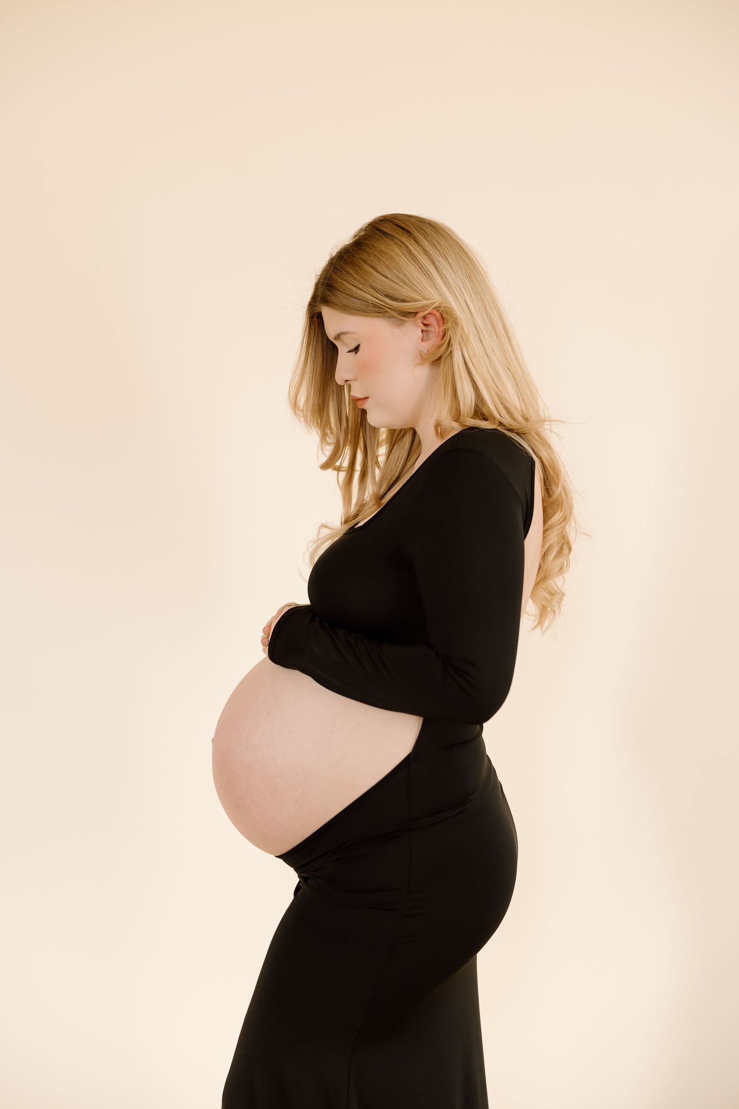 Side profile of an expectant mother in a black dress standing against a beige background.