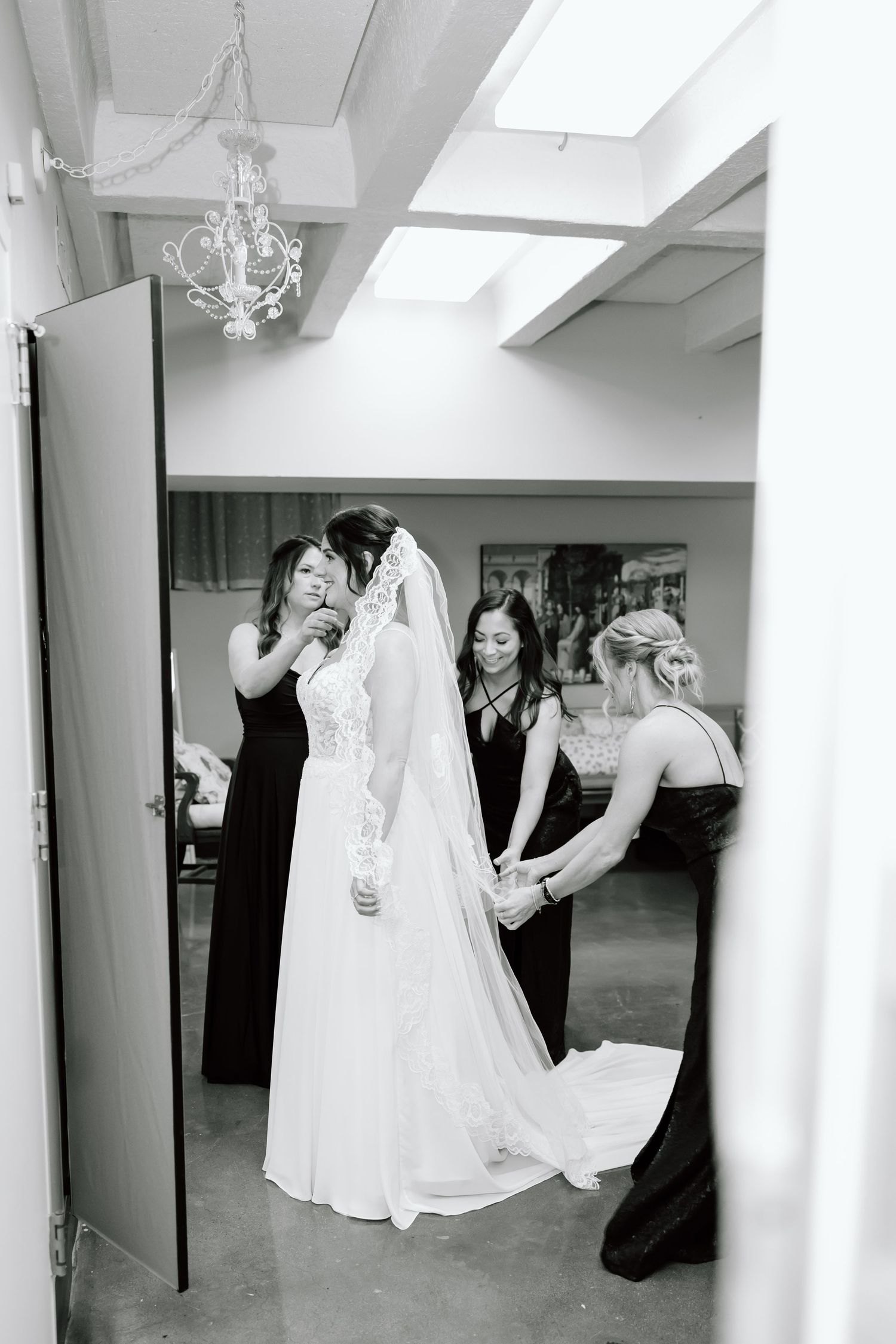 A bride in a white wedding gown gets final adjustments to her dress in a dressing room with a chandelier overhead.