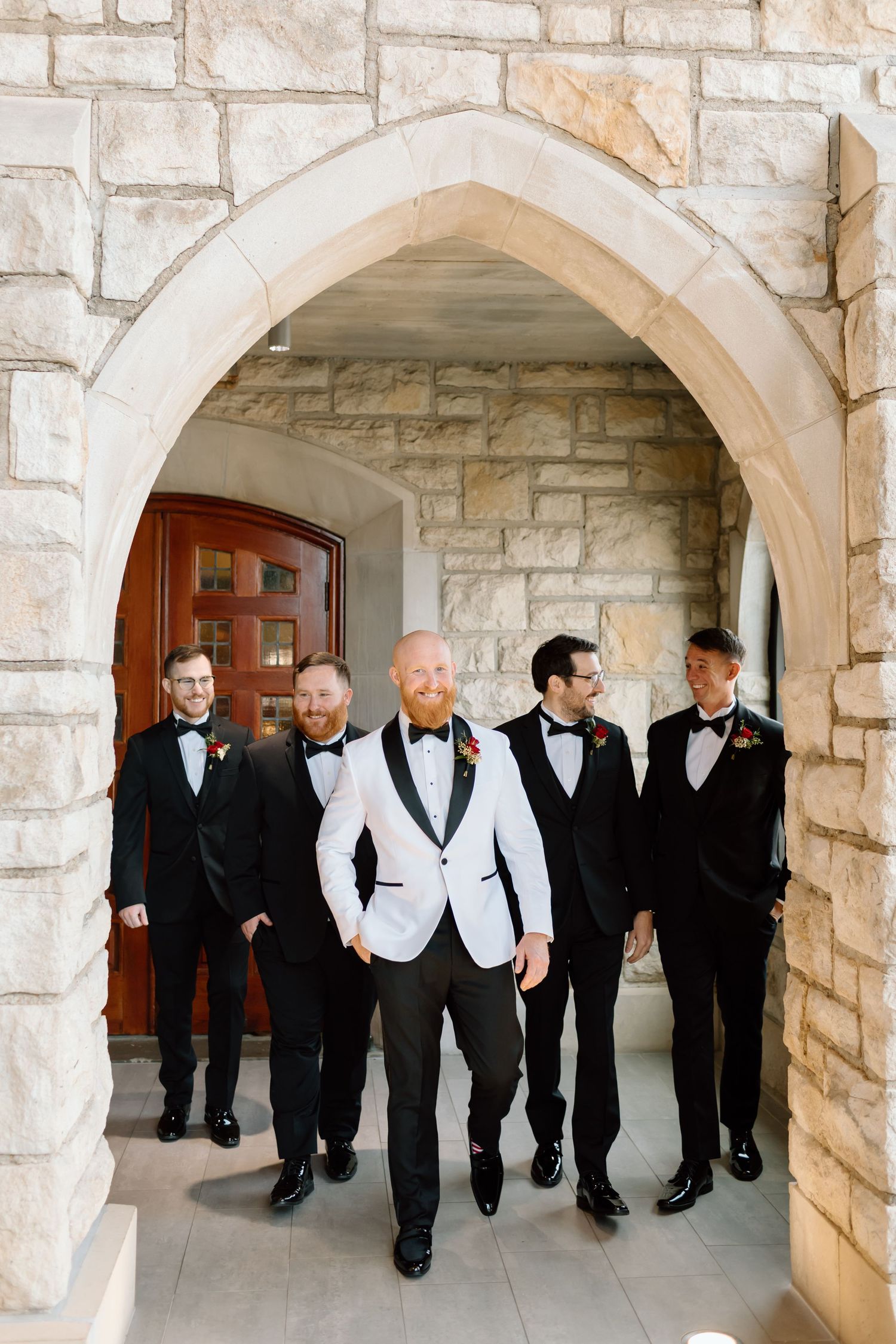 A group of groomsmen in formal black and white attire standing in an arched stone hallway.