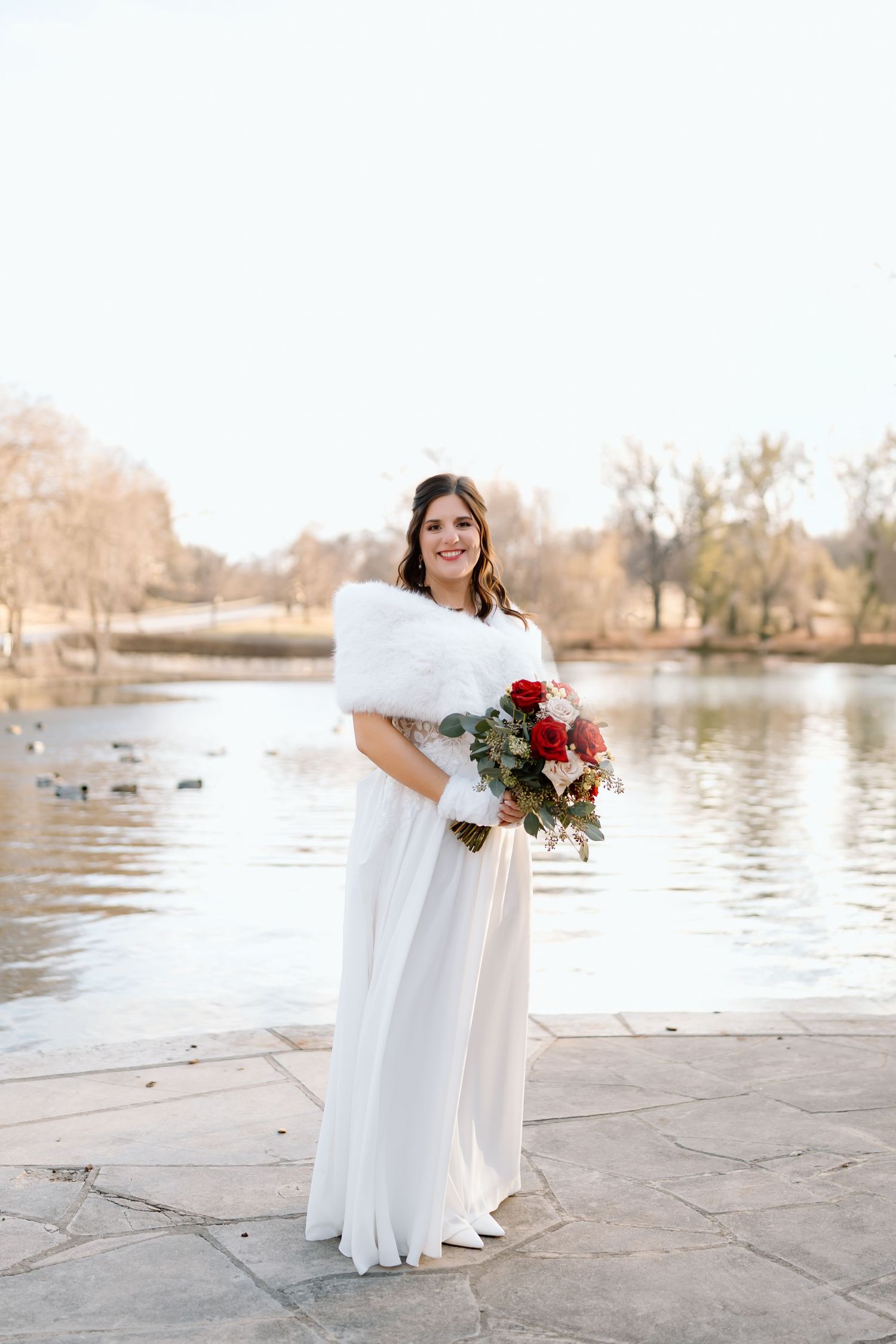 A bride in a flowing white dress holds a red and white bouquet while standing by a serene lake on a sunny day.