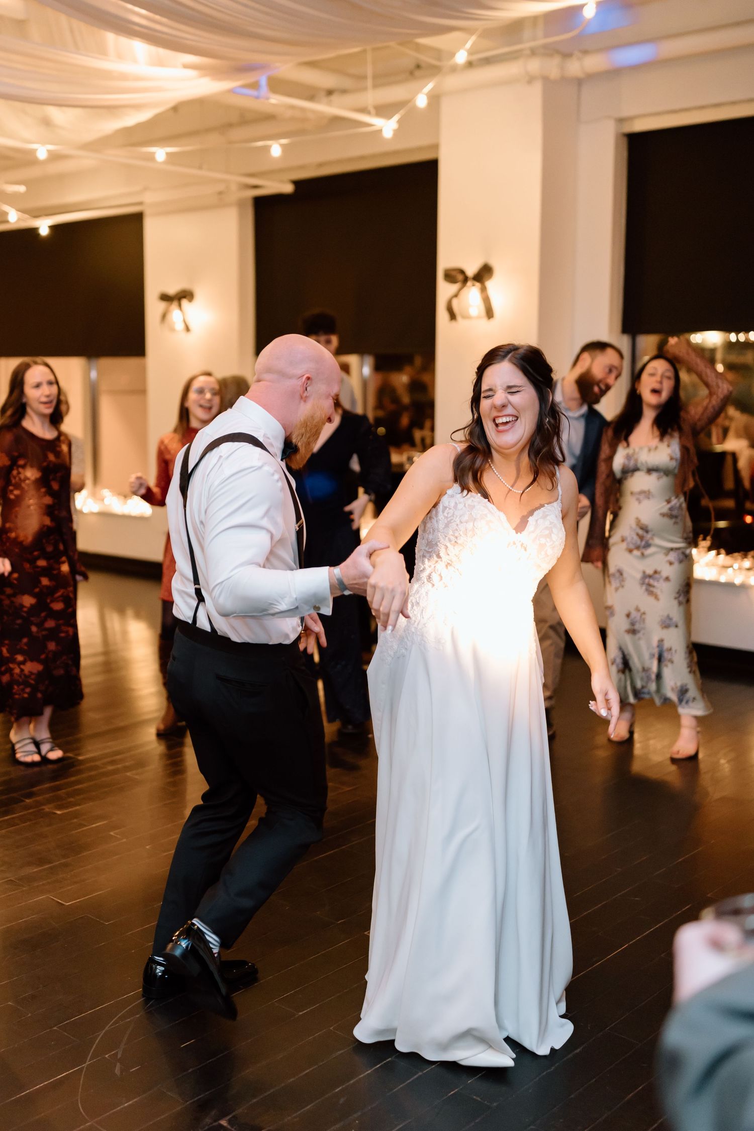 Wedding guests dance and celebrate together on a dark dance floor under white ceiling lights during an evening reception.