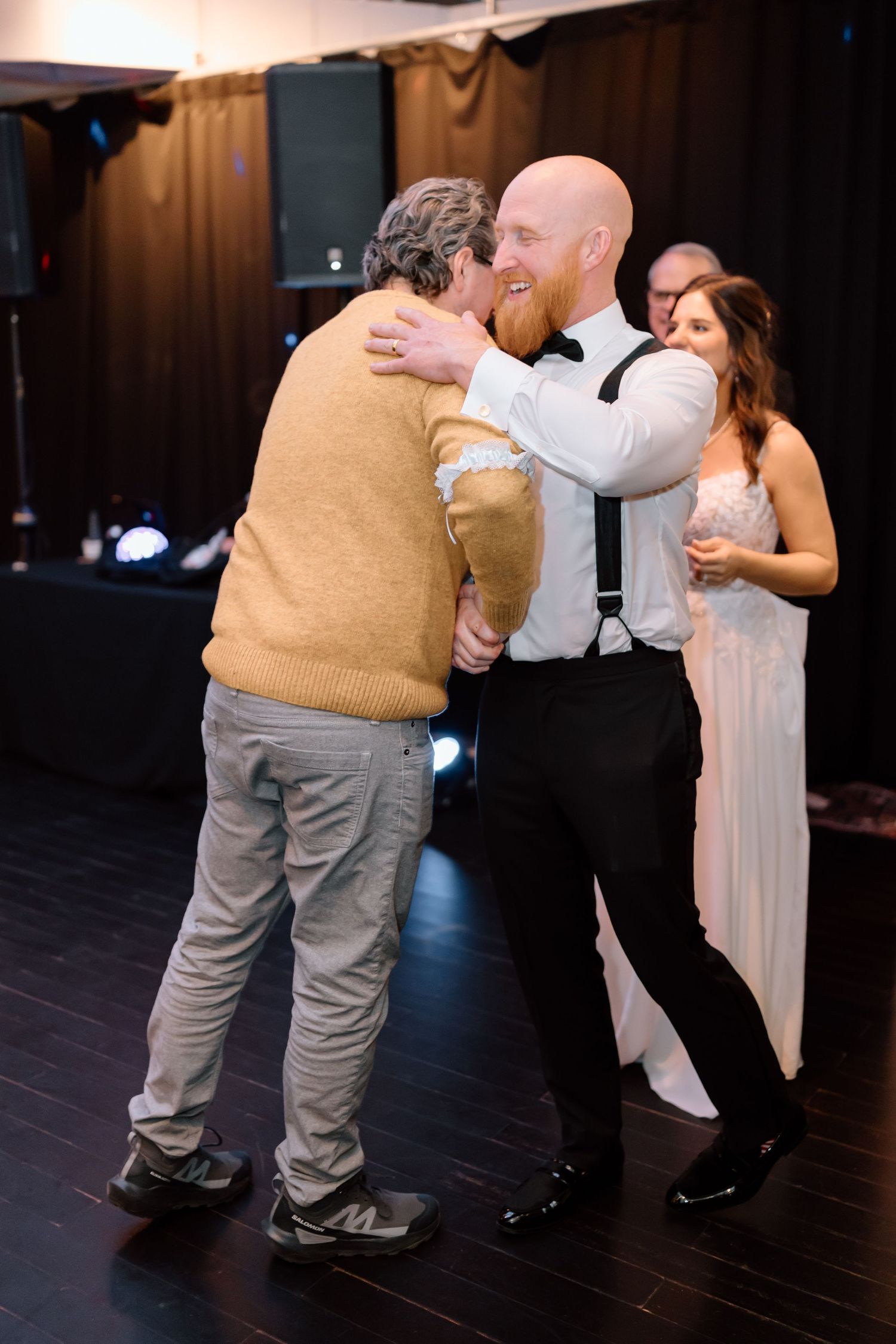 People embrace on a dark wooden dance floor at a wedding reception with one person wearing suspenders and a bowtie.