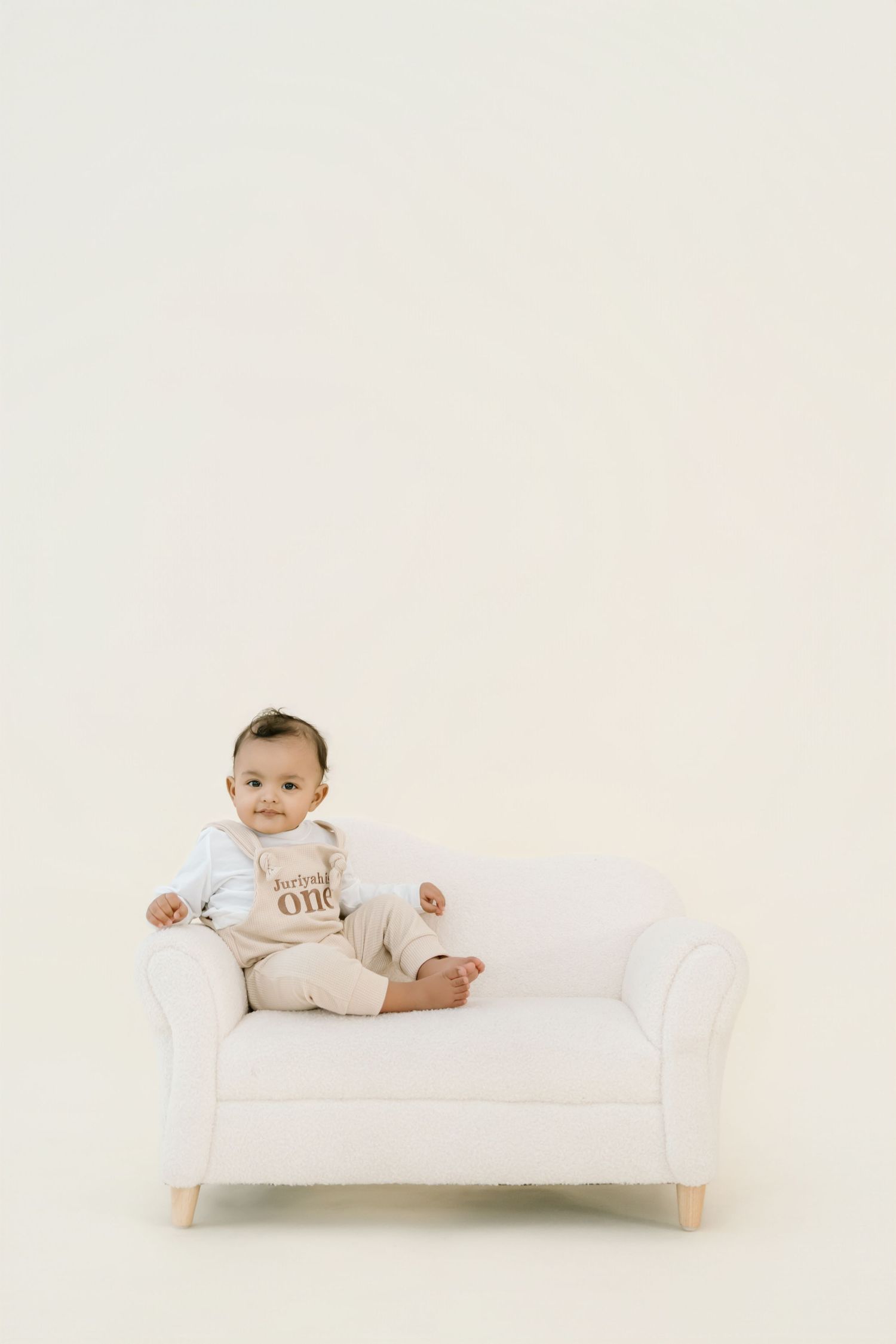 Baby in white onesie lying on minimalist white daybed against neutral background.