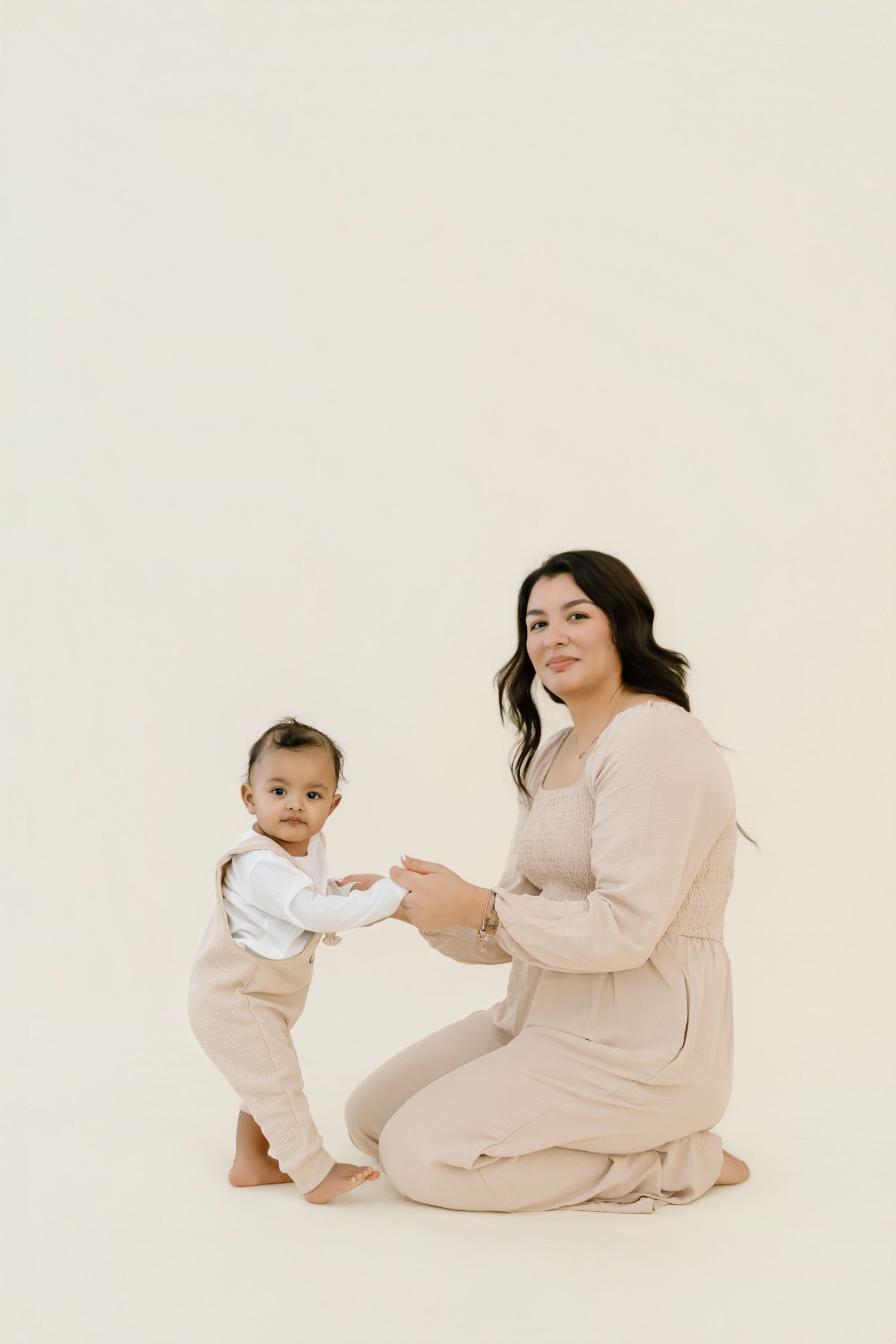 A parent and baby wearing matching cream-colored outfits sit and play together on a light beige background.