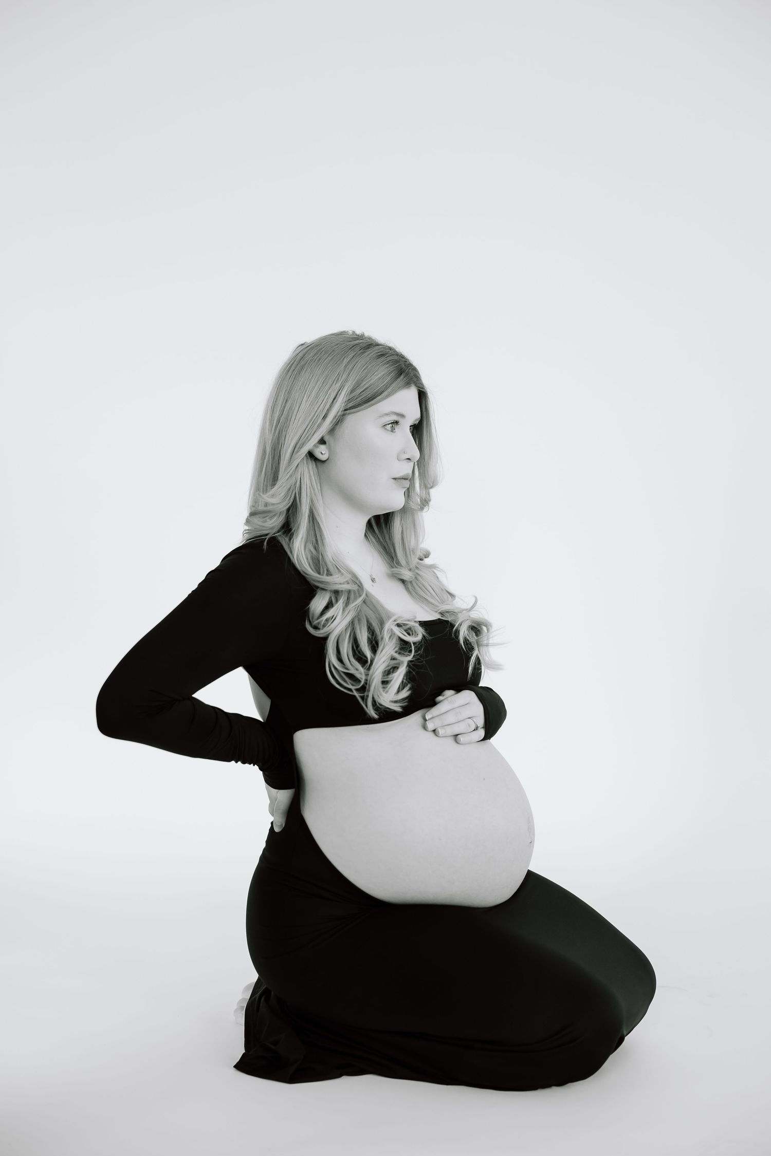 Professional maternity photo in black and white showing a woman in black dress kneeling on light background.