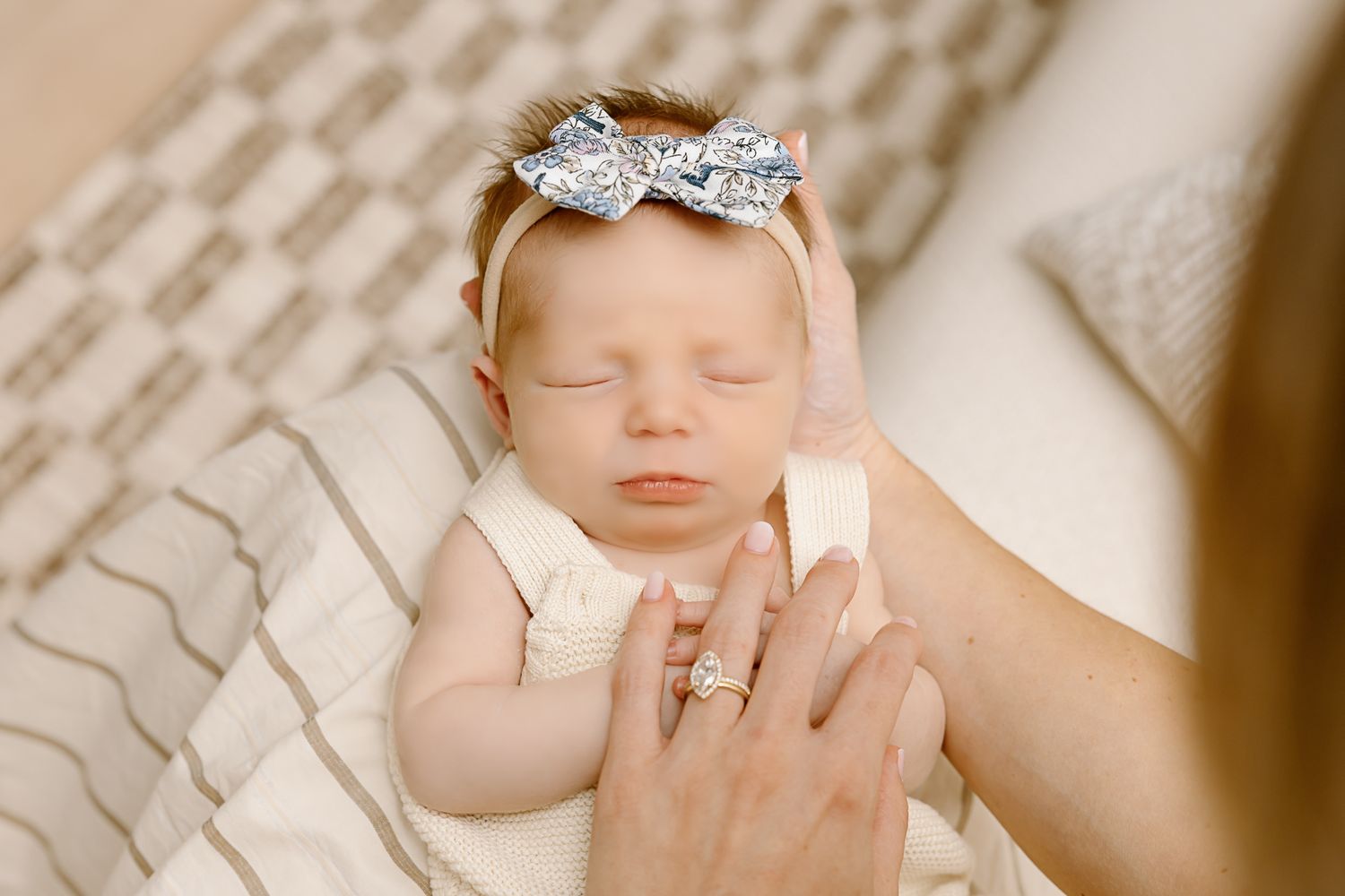 Sleeping newborn in cream romper with sparkly bow headband held by parent's hands.