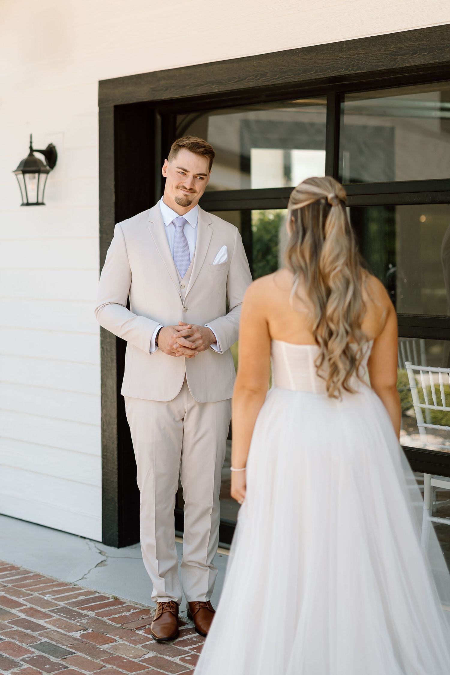 Groom in light beige suit awaiting first look on brick patio with white exterior.