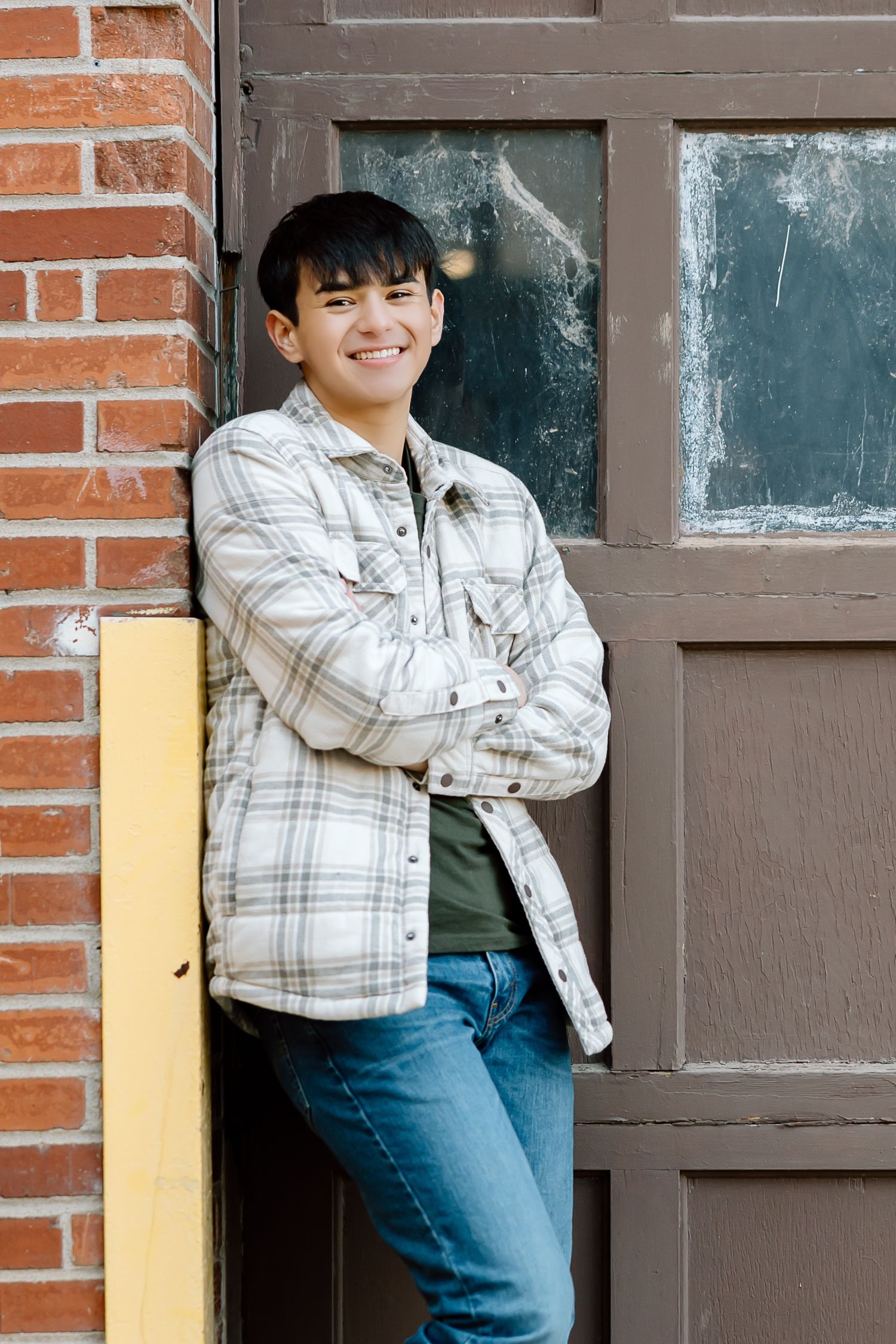 Person in plaid jacket smiling while leaning against brick wall and brown door.