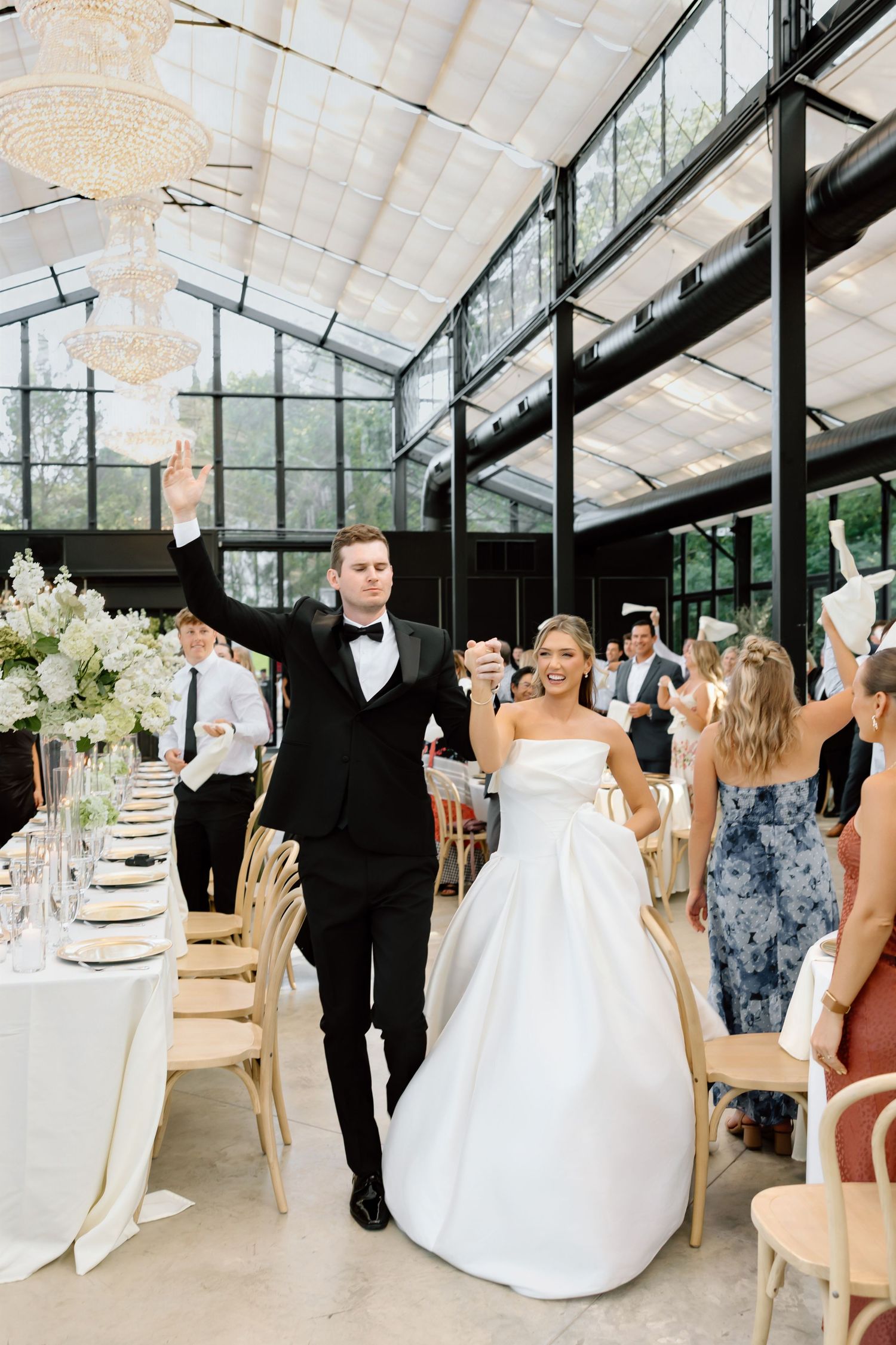 Bride and groom make grand entrance at elegant greenhouse wedding reception with chandeliers and floral arrangements.