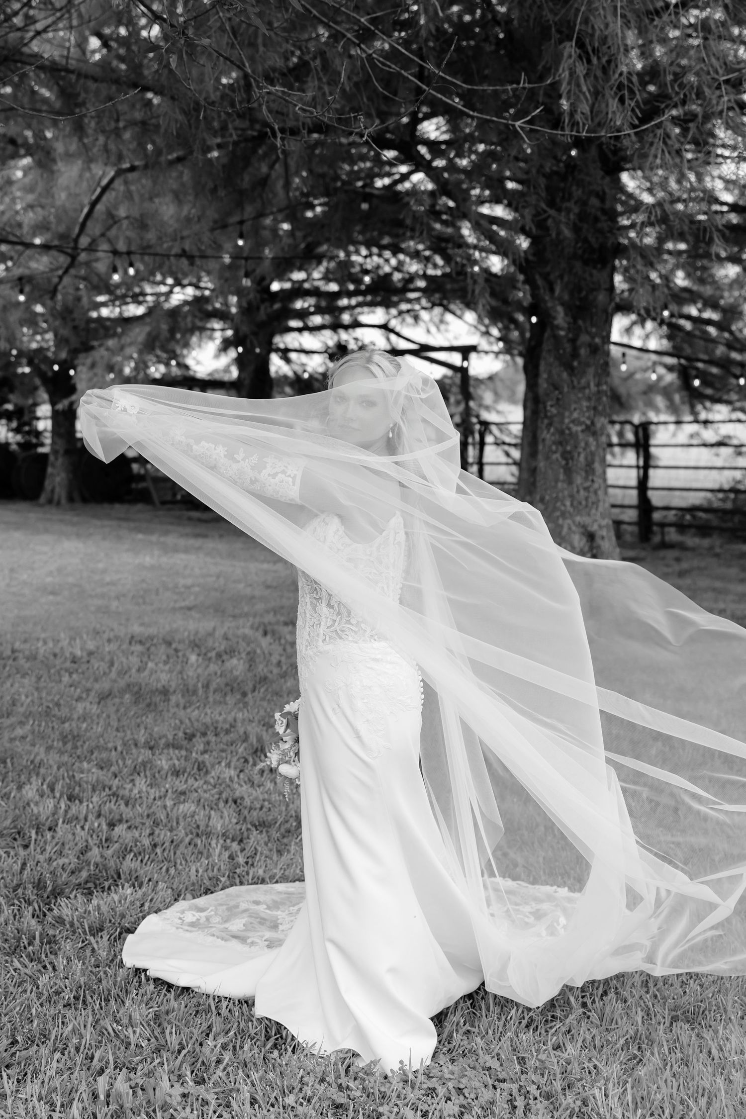 Black and white photo of a bride in a flowing gown with veil billowing in the wind outdoors near trees.