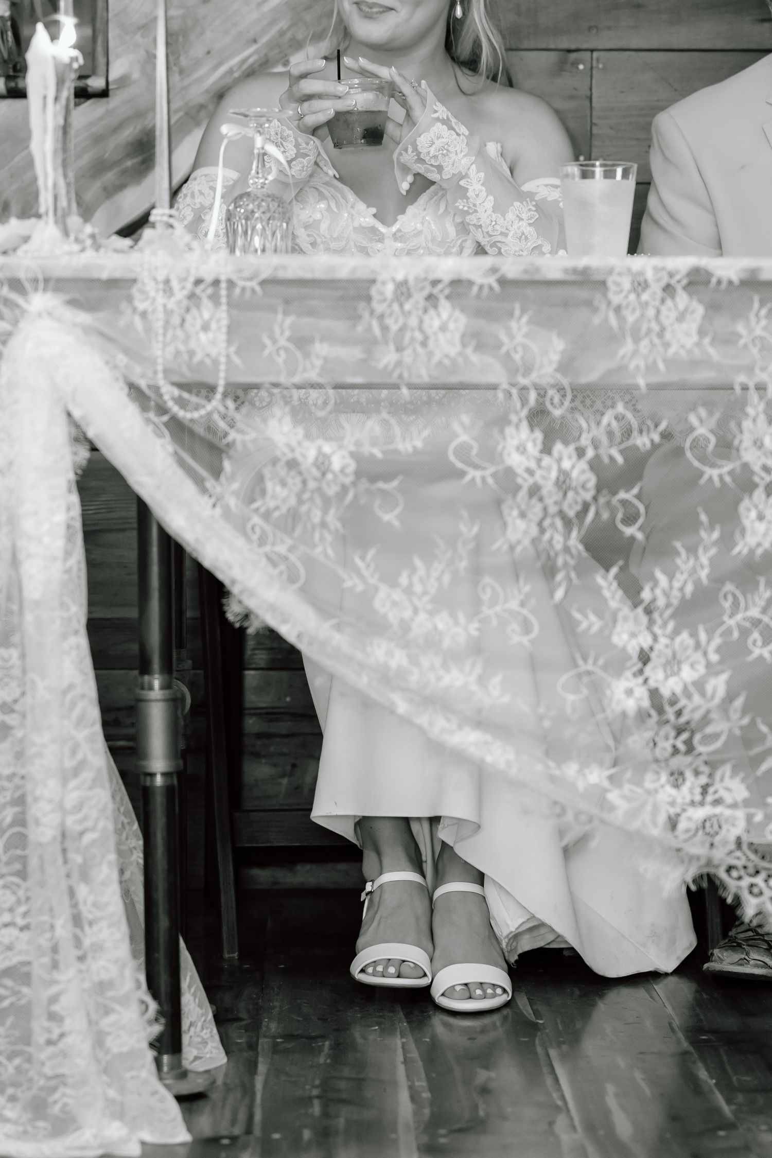 Black and white photo of a bride sitting at a lace-covered table, wearing heels and holding a drink at her wedding reception.