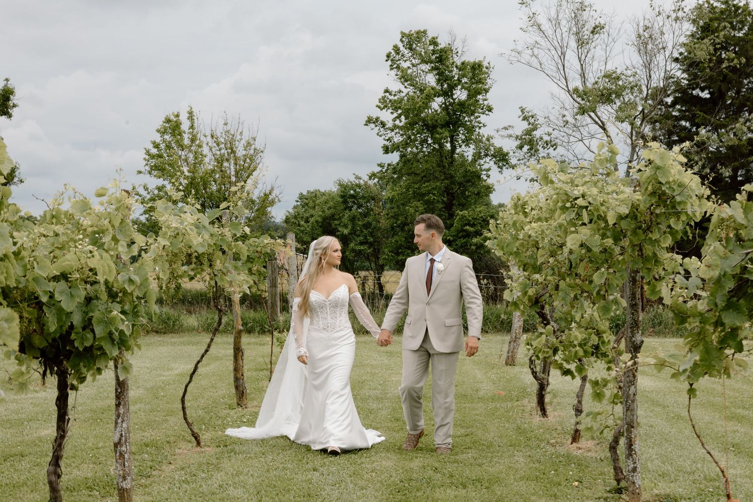 Bride and groom walk hand in hand through a lush vineyard on their wedding day under a cloudy sky.