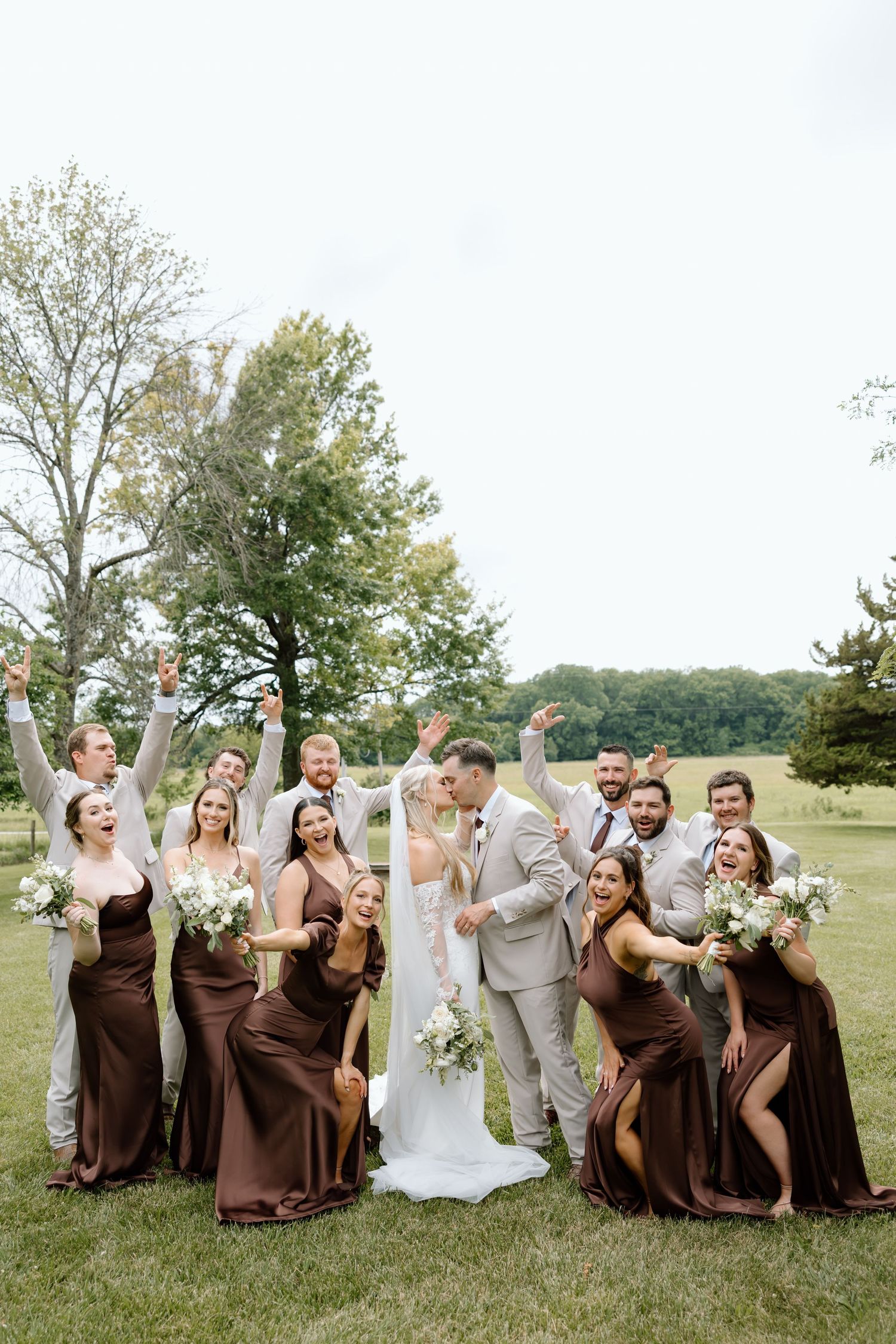 Wedding party celebrates outdoors, bridesmaids in brown gowns and groomsmen in tan suits cheering as couple kisses on green lawn.