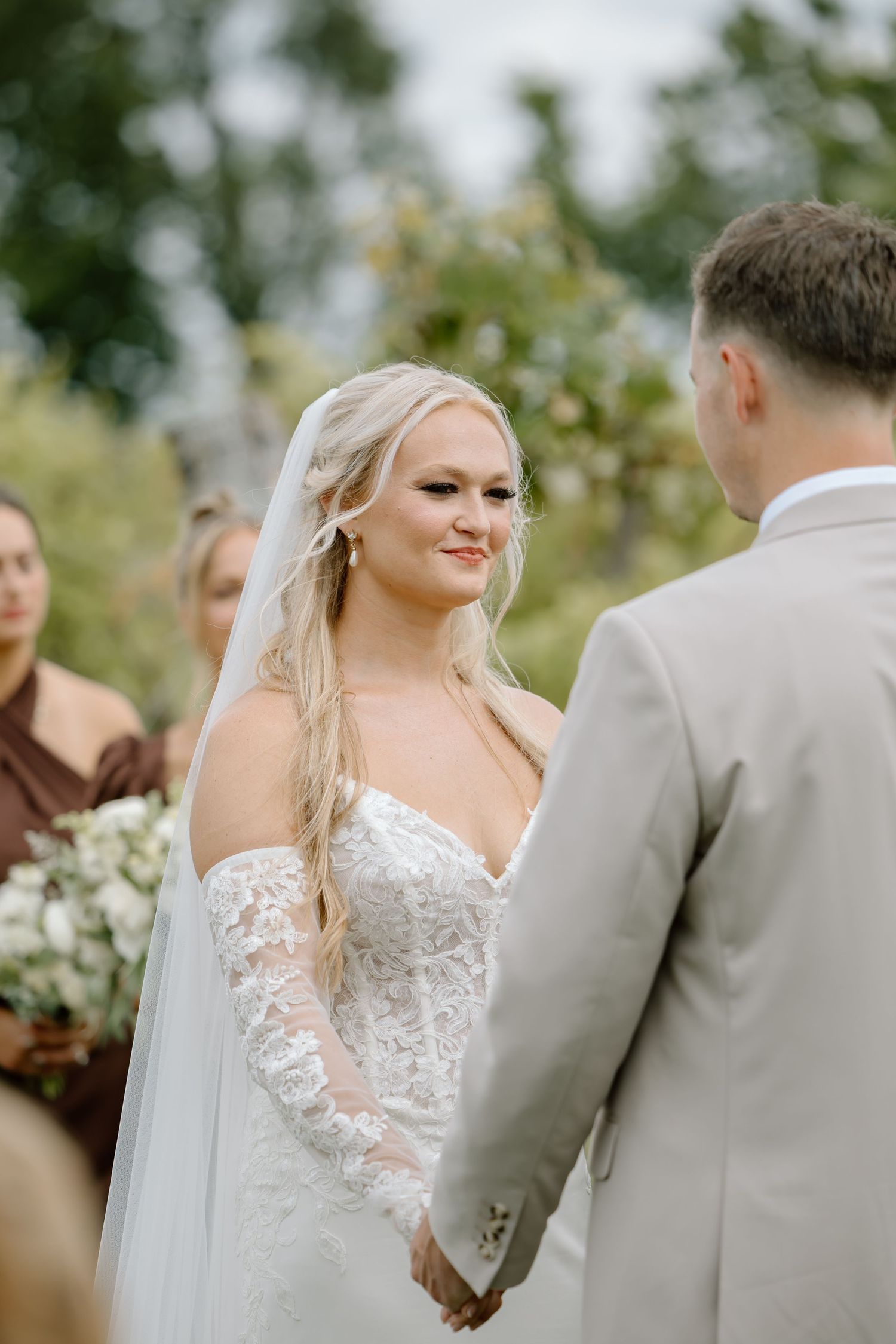 Bride in lace wedding gown and veil smiles at groom during outdoor wedding ceremony with bridesmaids in background.