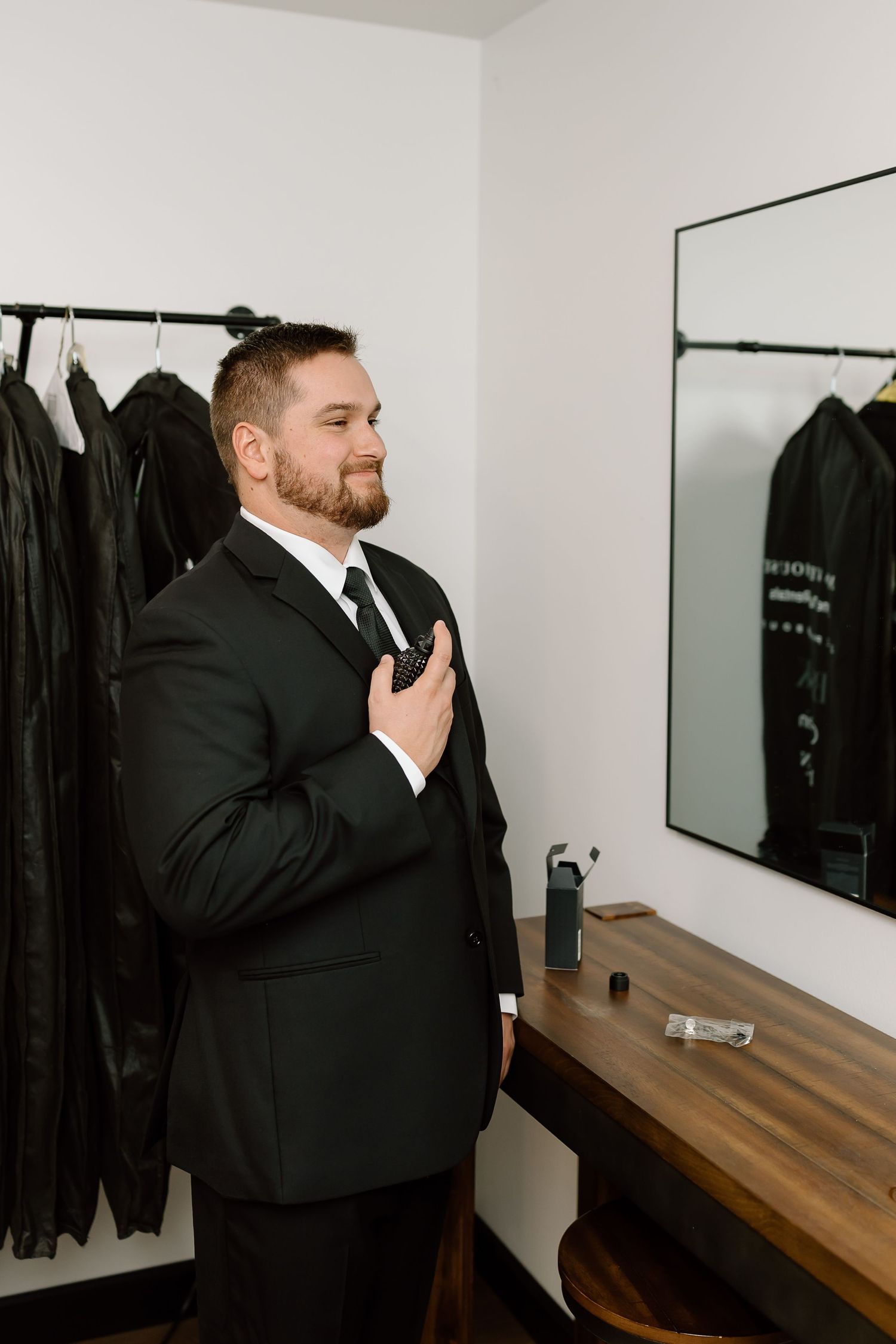 Man in black suit jacket stands near wooden counter and mirror in dressing room.