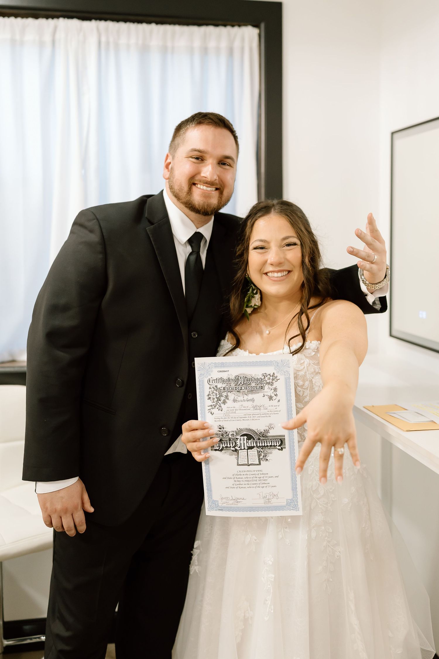 A couple in wedding attire shares a joyful moment while holding papers in a bright indoor setting.