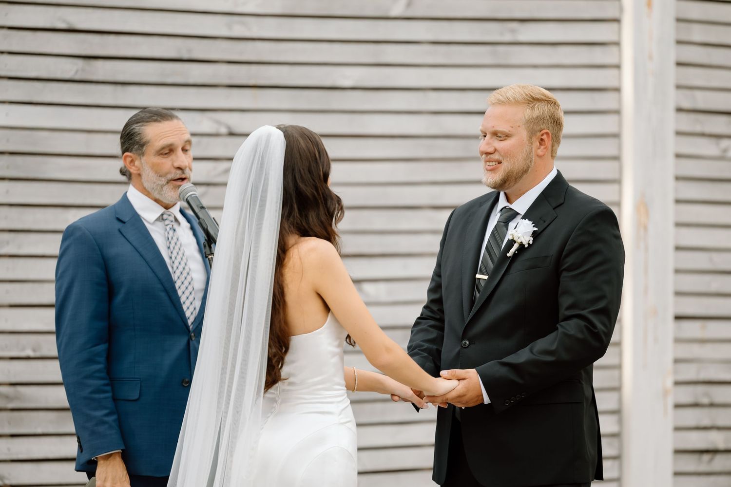 A couple shares a joyful moment during their outdoor wedding ceremony in front of a white wooden wall.