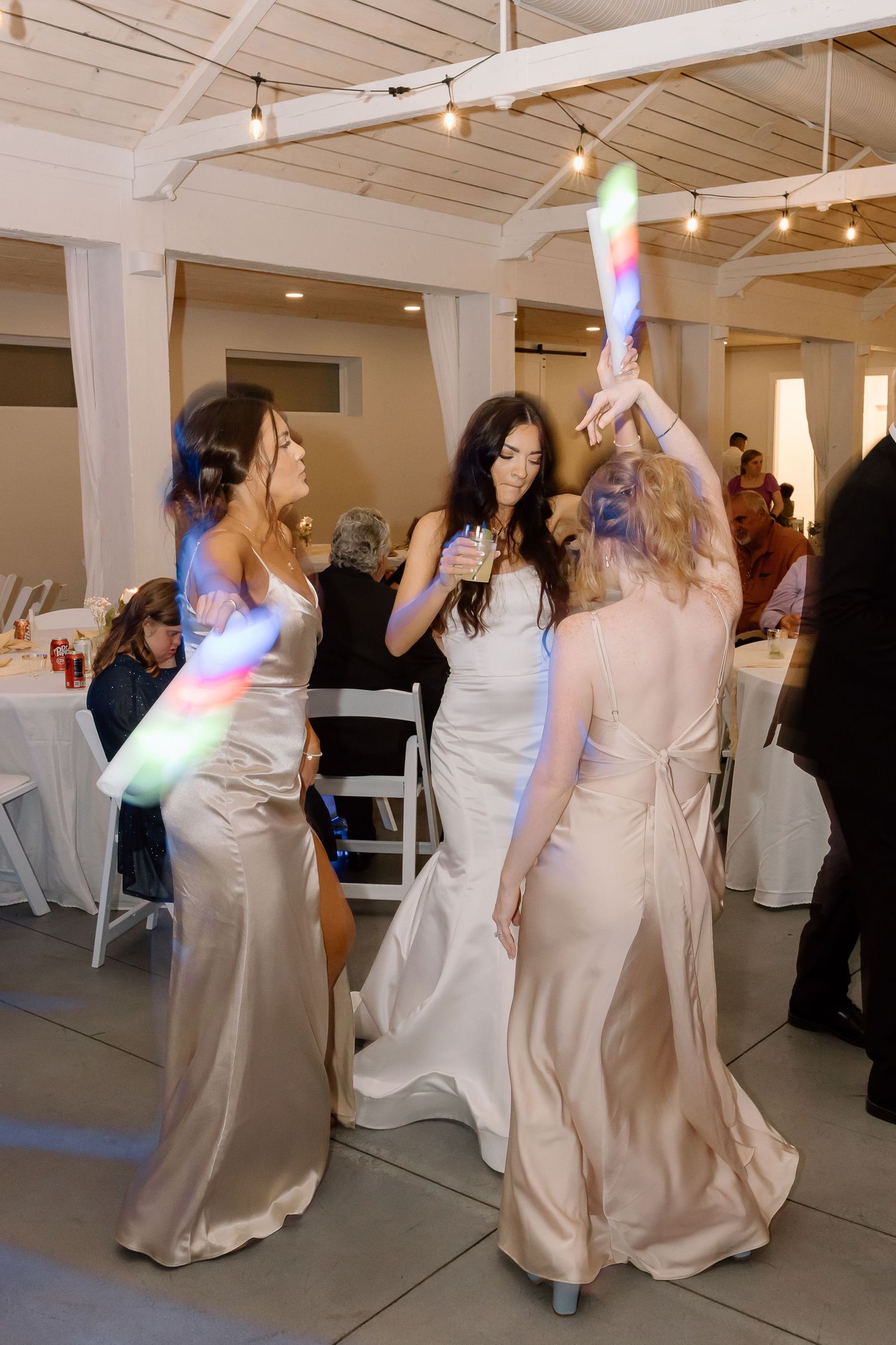 Wedding party guests dance and celebrate on a white-decorated indoor dance floor with colorful lights.