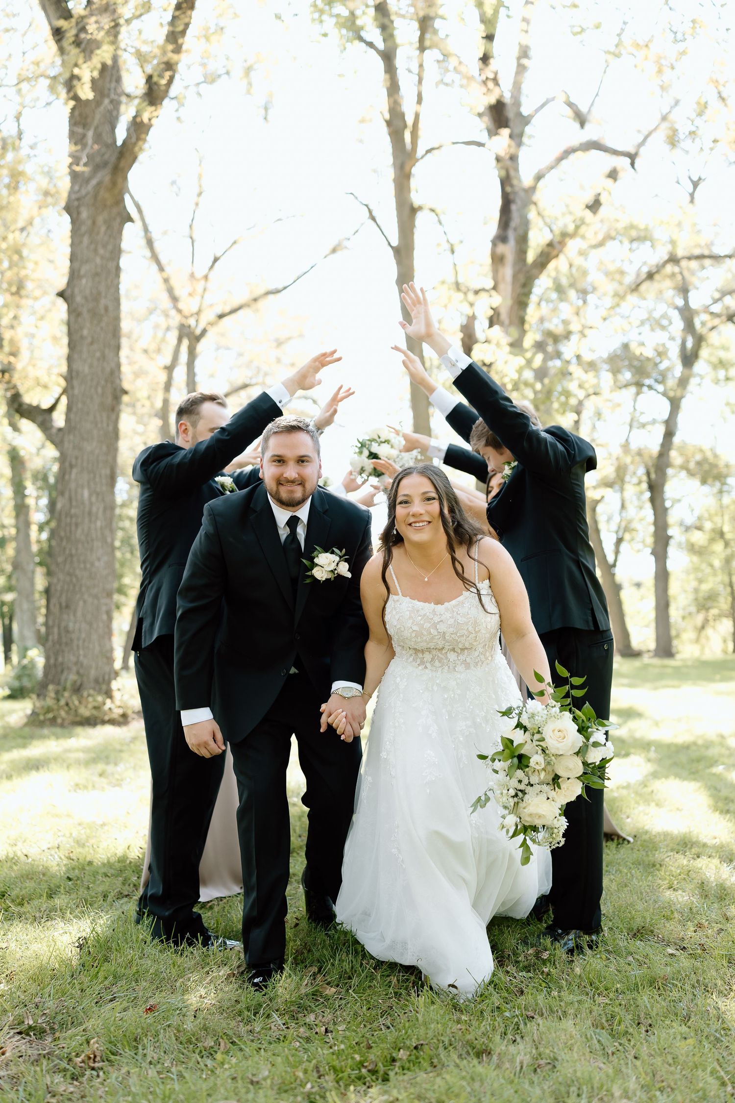 Wedding party in formal attire poses playfully in a wooded outdoor setting with natural sunlight filtering through trees.