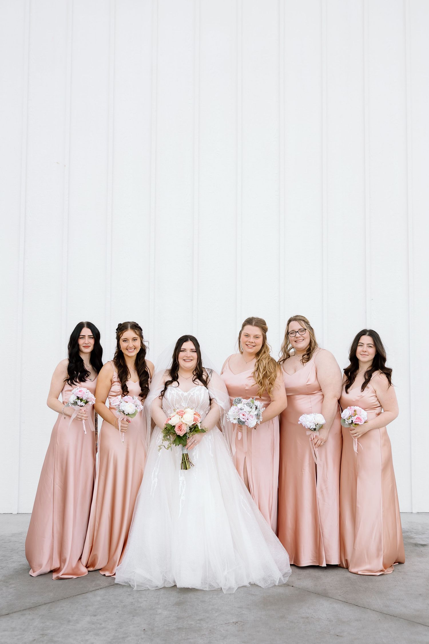 Bridesmaids in blush pink dresses stand together holding white bouquets next to bride in white gown.