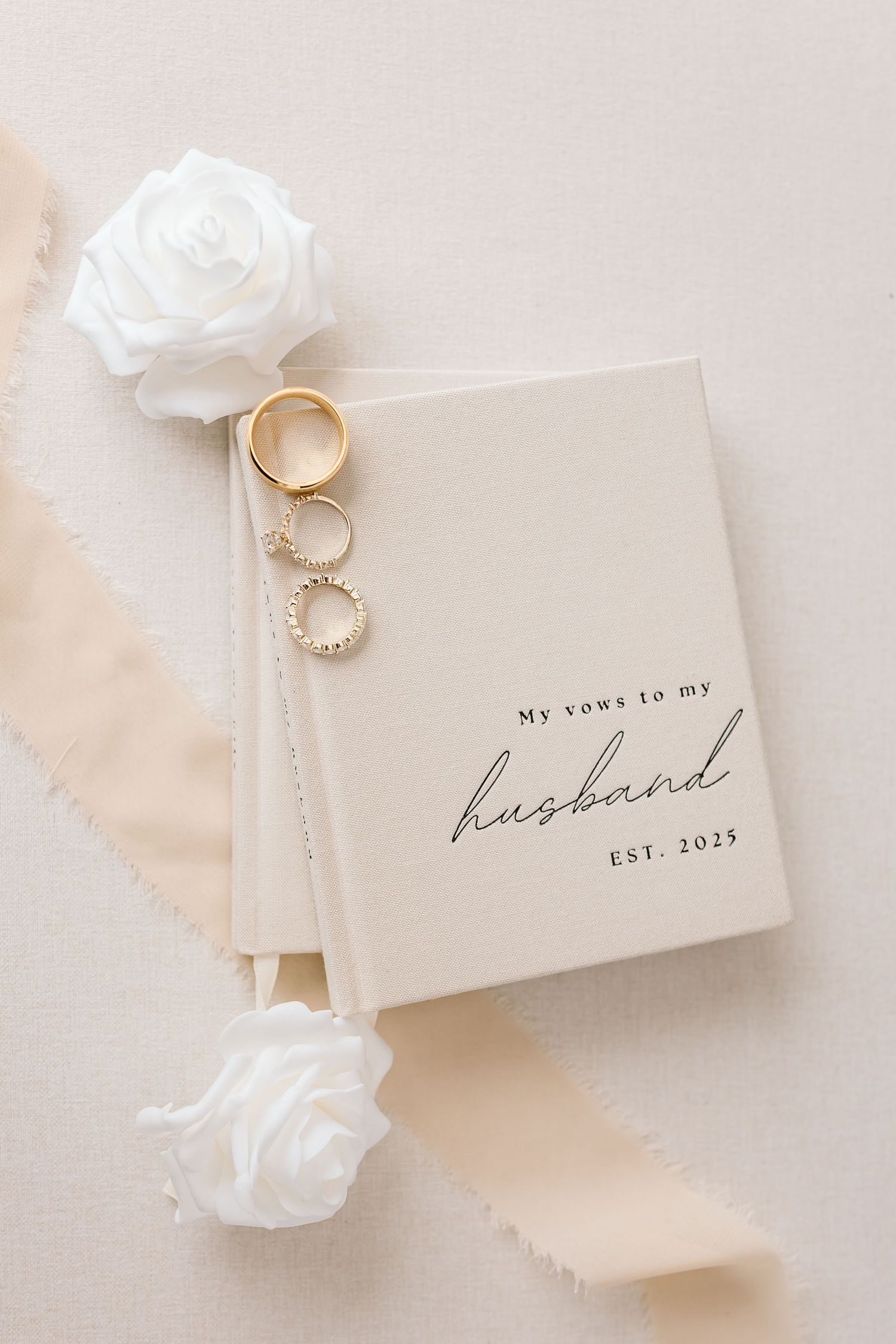 Wedding vow book with gold rings and white roses arranged on beige background in elegant minimalist style.