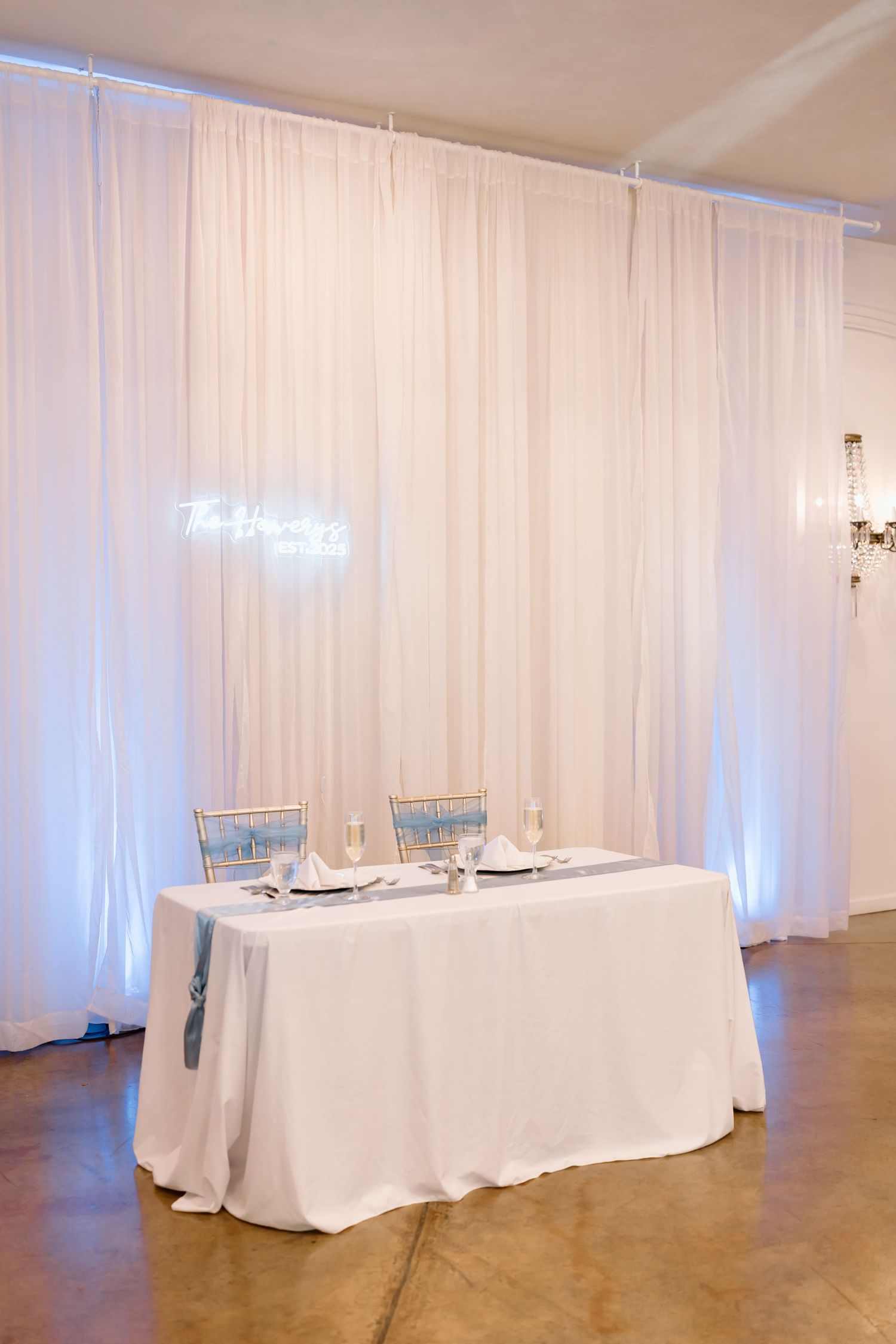 White draped curtains create an elegant backdrop for round dining tables in a modern event space with blue lighting.