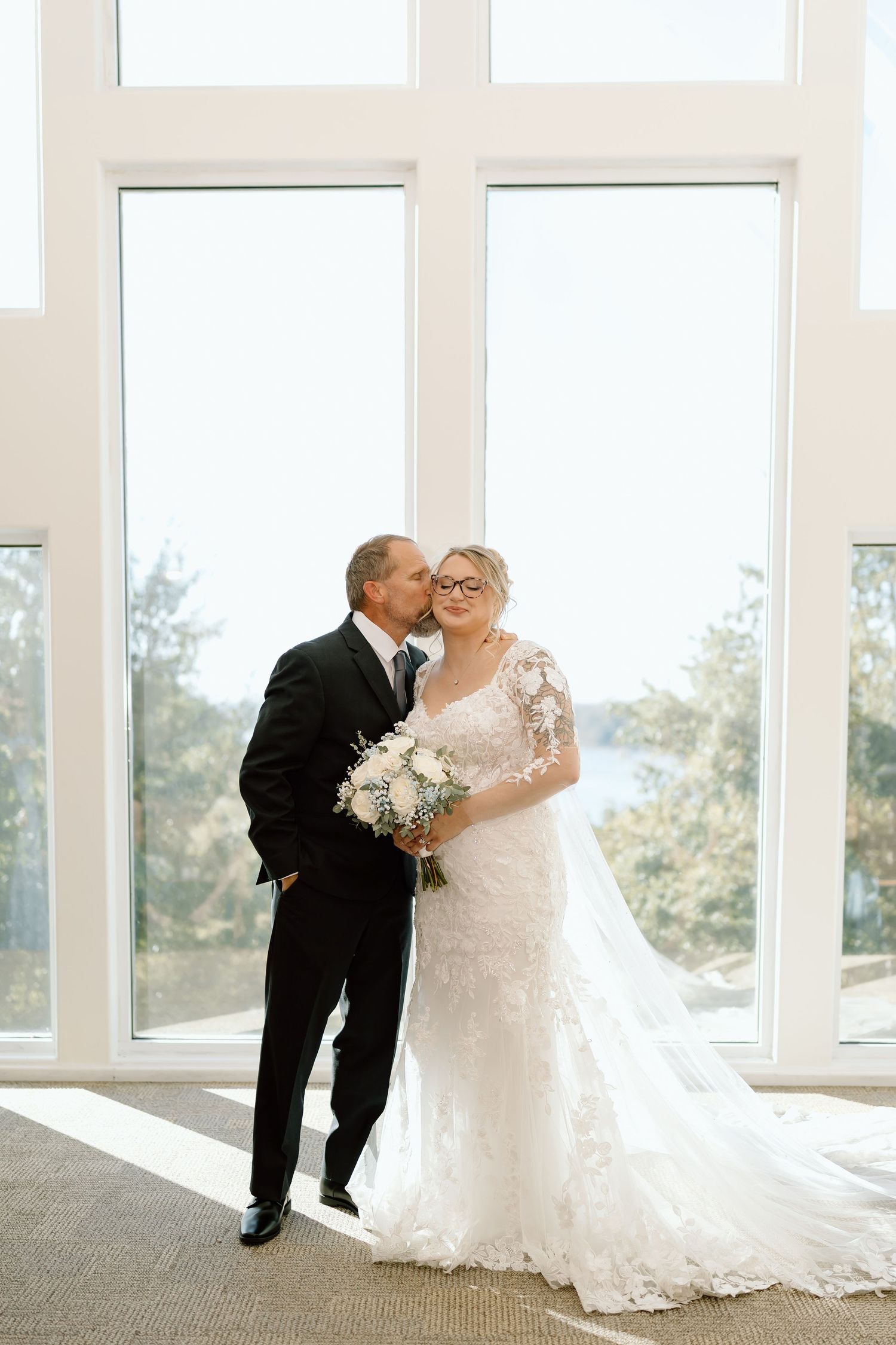 A couple shares an intimate moment by large windows on their wedding day, with the bride in a lace gown holding white flowers.