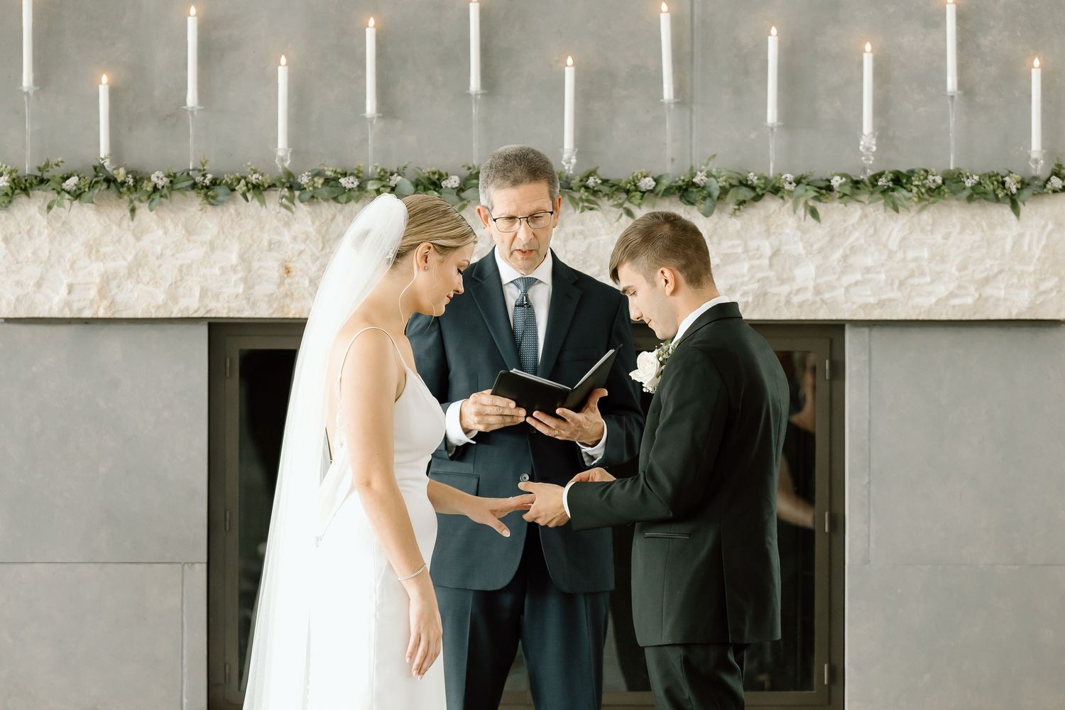 Wedding couple stands with officiant during intimate outdoor ceremony with white picket fence in background.