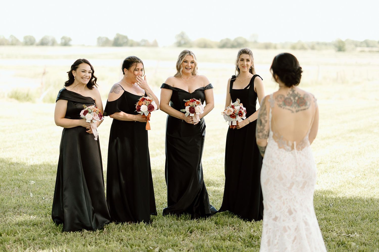 A wedding party in black dresses and a white gown laughs joyfully together in a sun-lit outdoor setting.