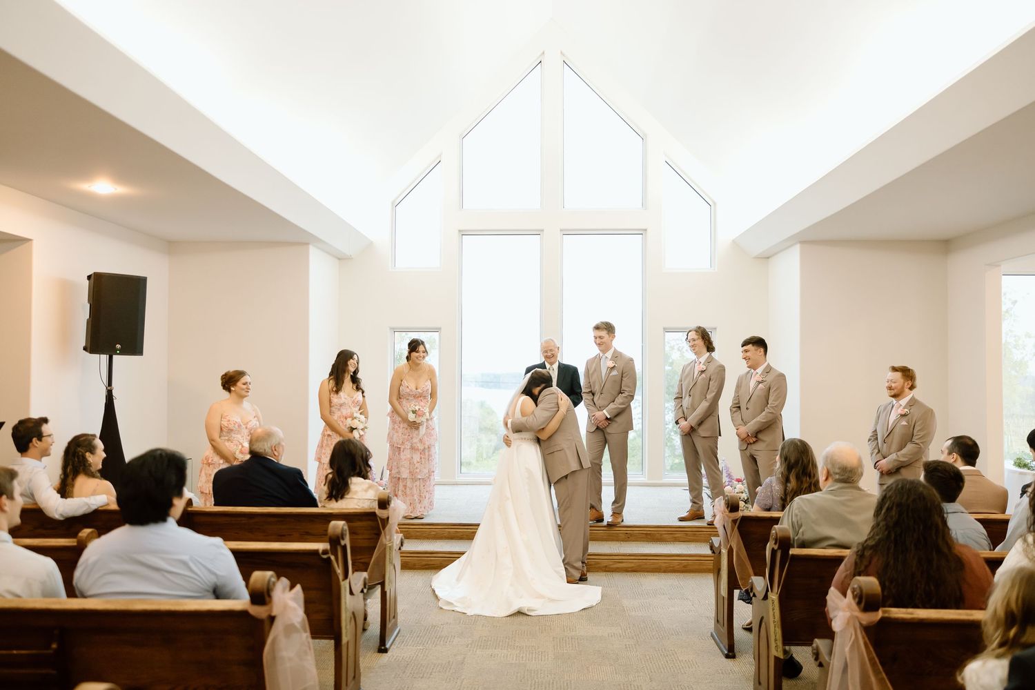 Wedding ceremony guests sit in wooden pews inside a bright white minimalist church with natural light streaming through windows.