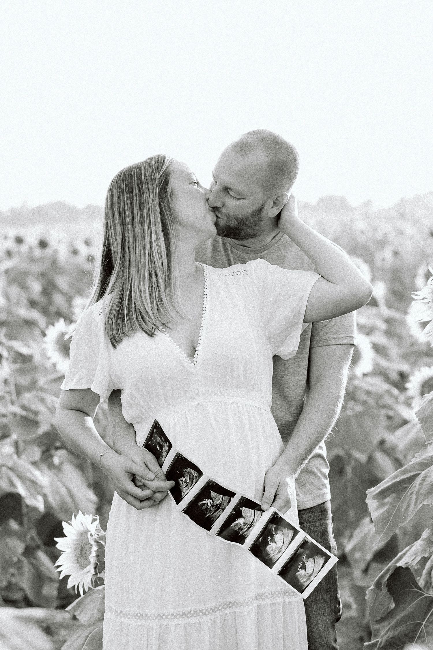 A couple in white clothing shares tender moments in a sunflower field while holding ultrasound photos.